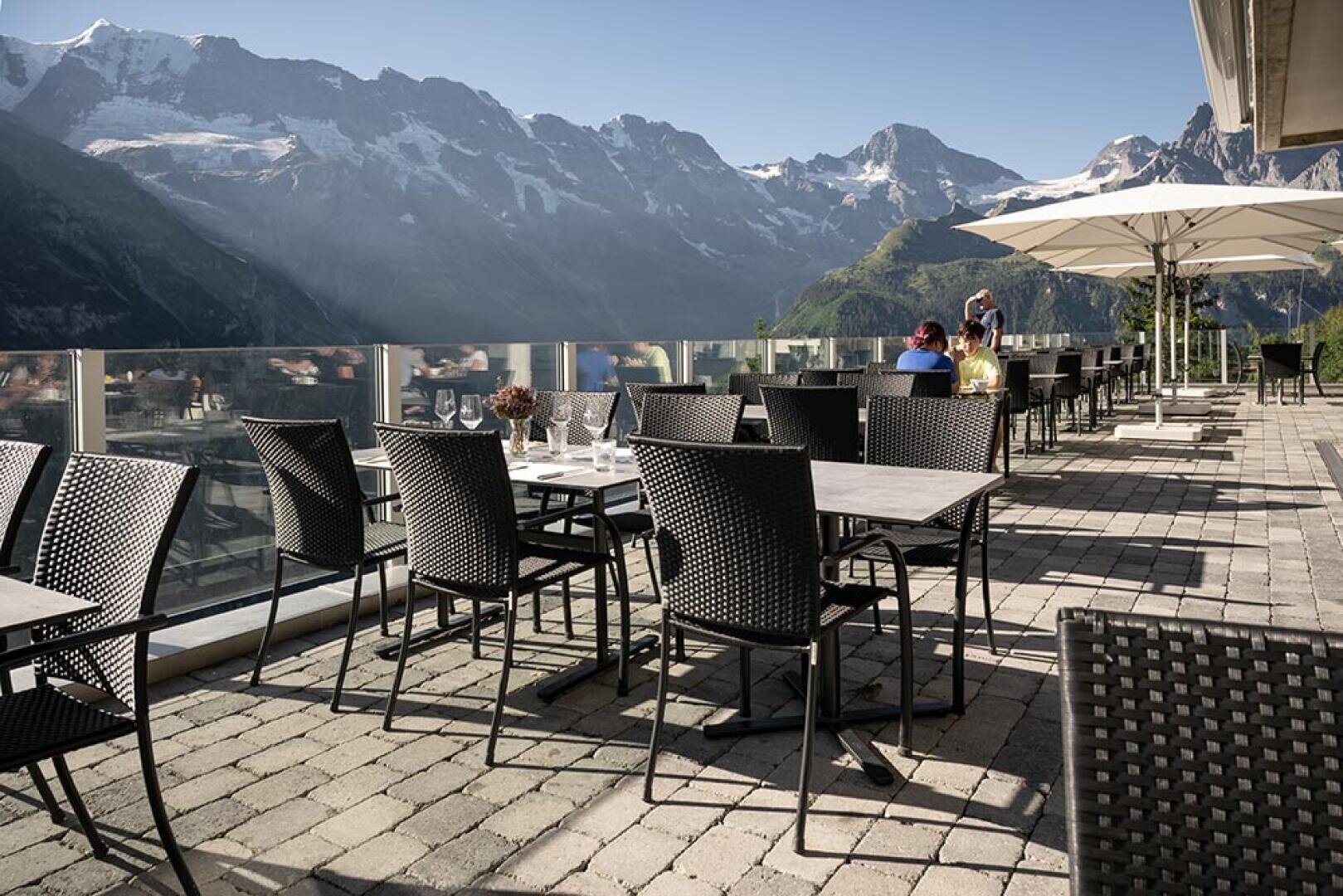 Restaurantterrasse mit schwarzen Korbstühlen und Tischen, Blick auf eine malerische Bergkette mit schneebedeckten Gipfeln unter blauem Himmel; ein paar Leute sitzen unter großen weißen Sonnenschirmen.