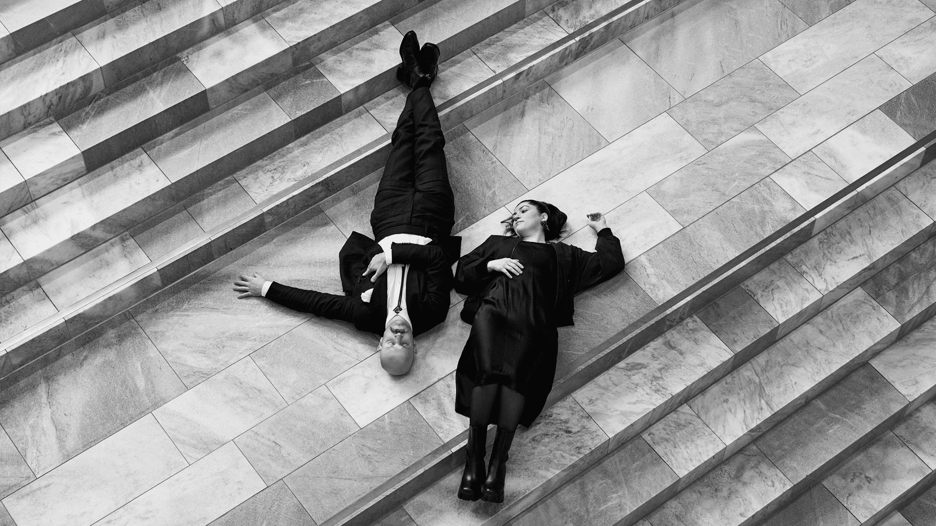 Ein Mann im Anzug und eine Frau im Kleid liegen nebeneinander auf einer gefliesten Treppe, beide sehen entspannt aus. Das Bild ist schwarz-weiß, von oben gesehen, mit geometrischen Stufen, die diagonale Linien über die Szene bilden.