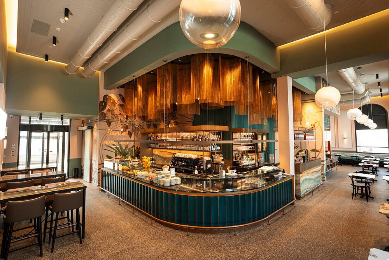 Café moderne et élégant avec un bar central doté de panneaux sarcelle, de touches dorées et de décorations métalliques suspendues. L'espace est doté de grandes lampes globe, de hauts plafonds et de sièges confortables, ce qui crée une atmosphère chaleureuse et accueillante.