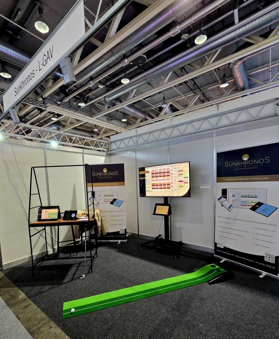 Le stand de Sunkhronos se compose de bannières, d'une table avec des appareils électroniques, d'un grand écran affichant des données et d'un mini putting green avec une balle de golf. Le stand est installé dans un hall d'exposition.