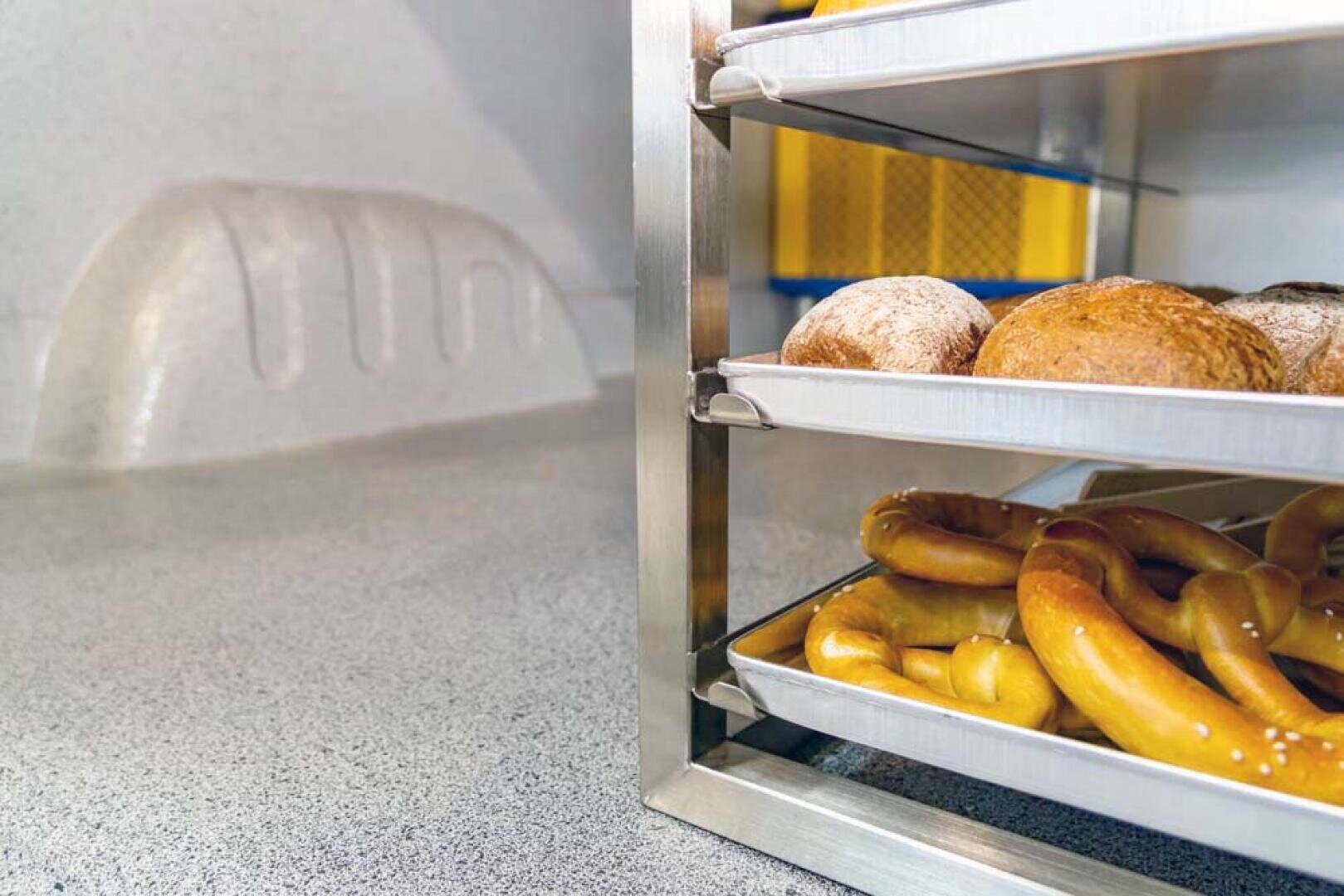 Trois plateaux métalliques contenant des produits de boulangerie, dont des petits pains et des bretzels, sont empilés sur un support dans une cuisine ou une boulangerie avec un plan de travail texturé et un arrière-plan industriel.