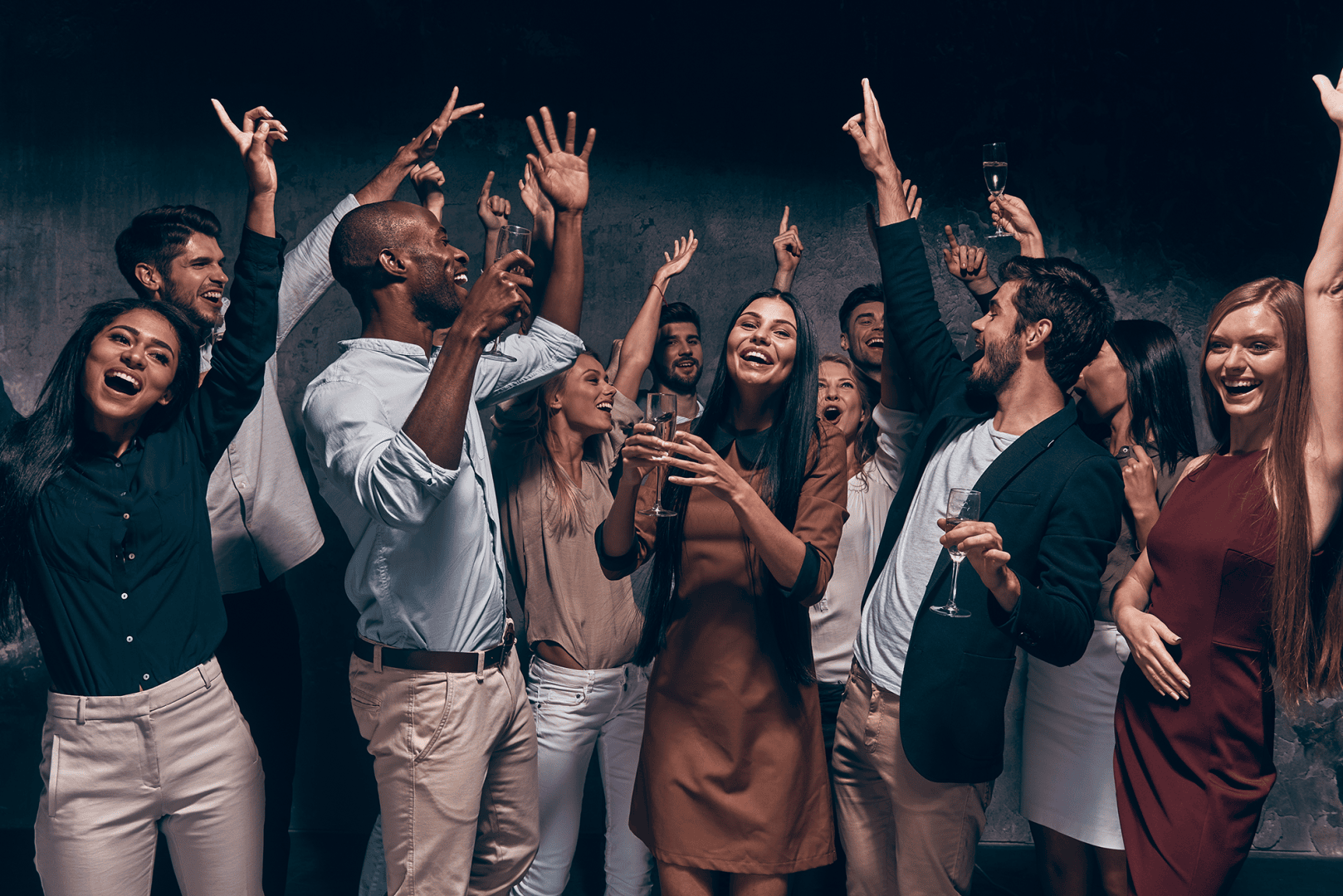 Eine Gruppe junger Erwachsener in halb-formeller Kleidung feiert gemeinsam auf einer Party, lächelt und hebt vor einem dunklen Hintergrund aufgeregt ihre Arme und Gläser.