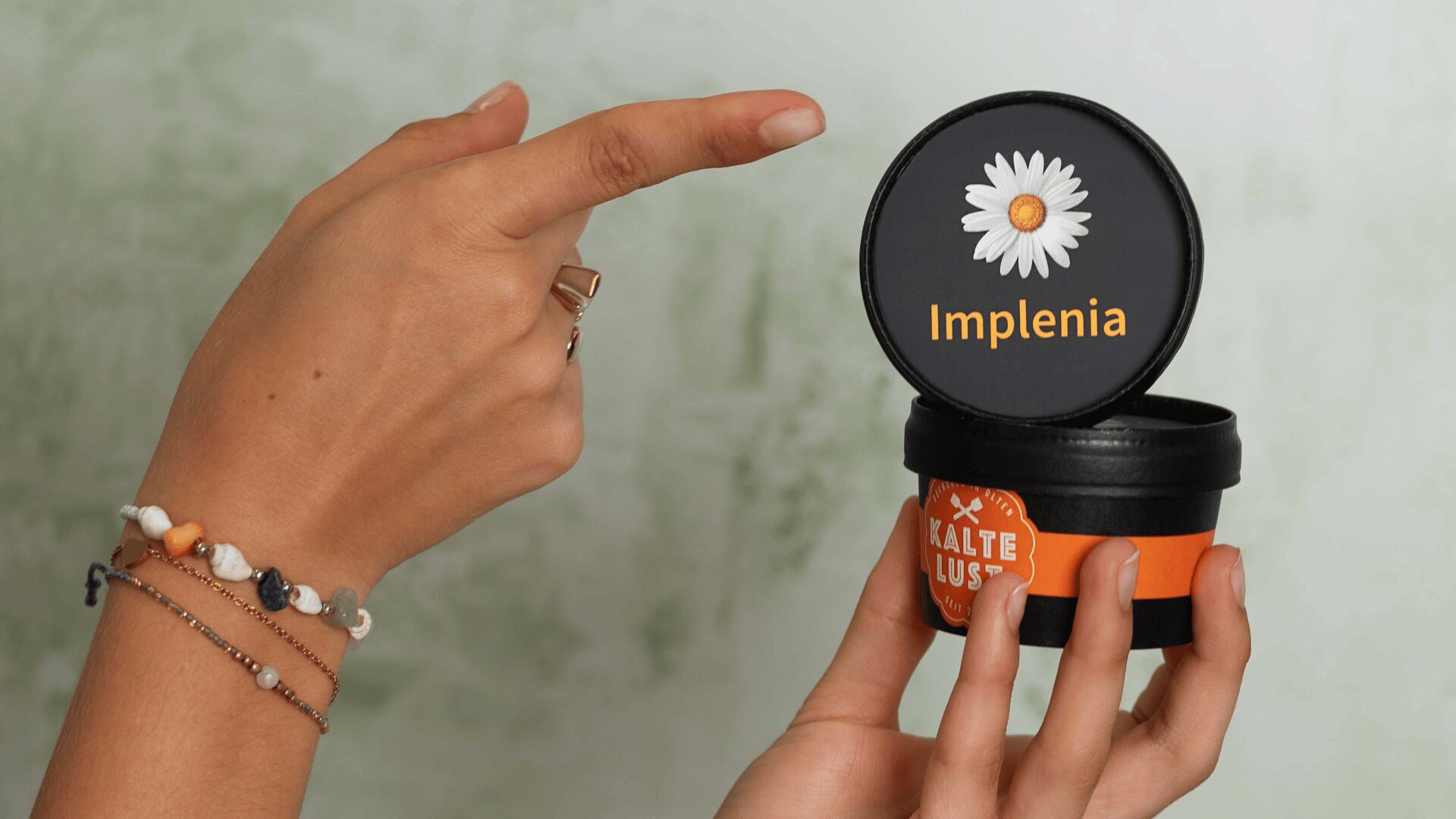 Eine Person hält einen kleinen schwarzen Behälter mit einem orangefarbenen Etikett in der Hand und hebt den Deckel an, auf dem ein Gänseblümchen und die Aufschrift Implenia zu sehen sind. Die Person trägt Armbänder und einen Ring.