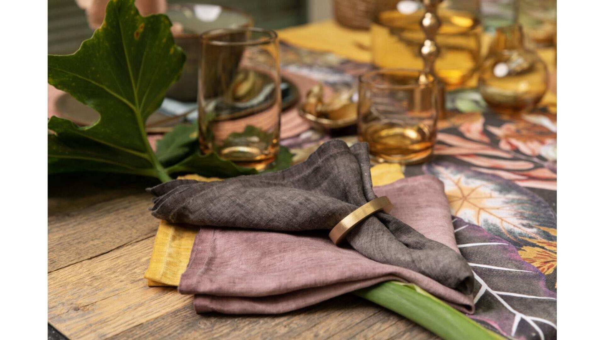 Une table rustique en bois est ornée d'une feuille tropicale, de serviettes de table en lin aux couleurs discrètes, d'un rond de serviette en or, de verres ambrés et d'un chemin de table floral coloré, créant ainsi une atmosphère chaleureuse et élégante.