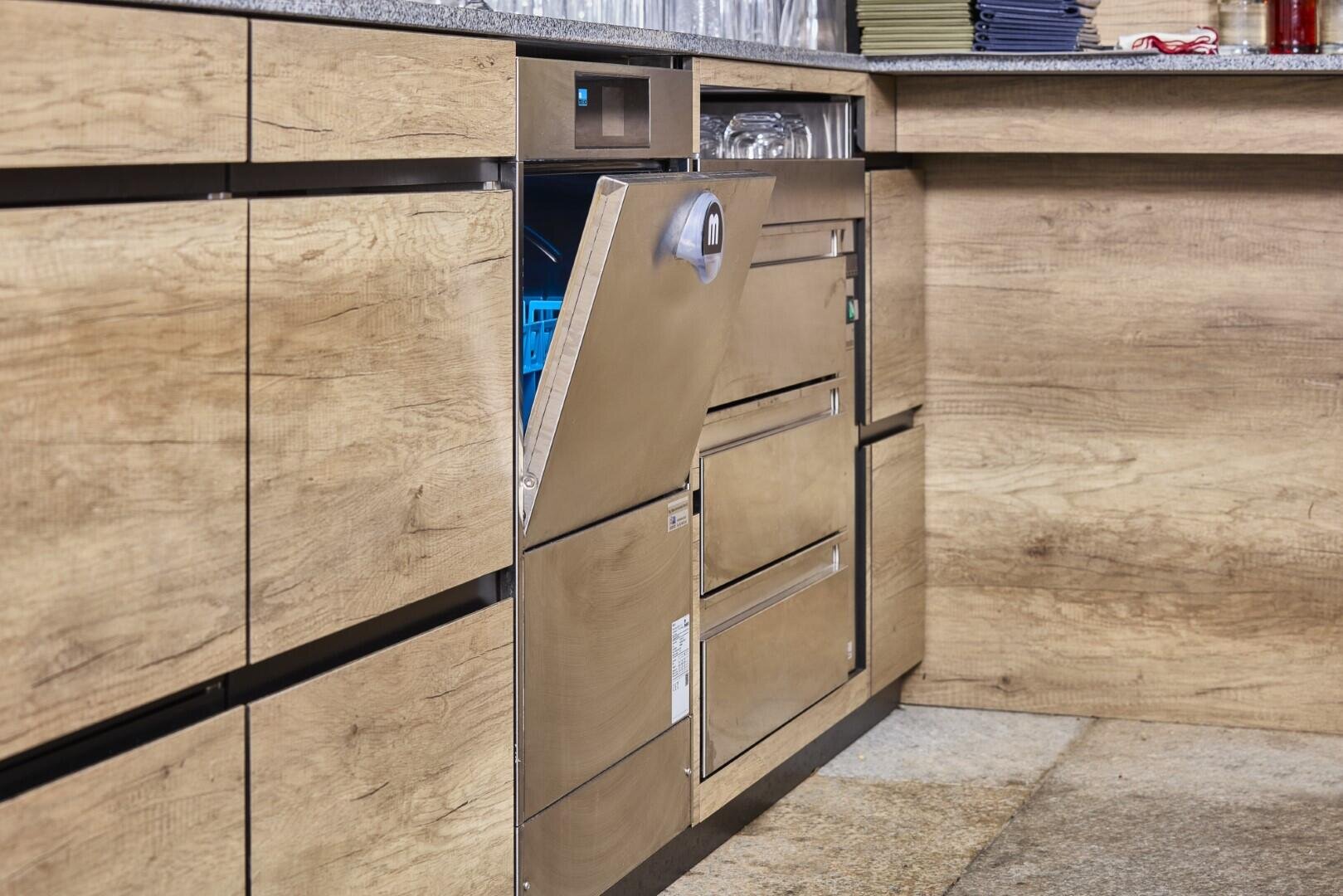 Ein teilweise offener Geschirrspüler aus Edelstahl ist in die hölzernen Küchenschränke eingebaut, wobei das saubere Geschirr im Inneren sichtbar ist. Auf der Arbeitsplatte stehen Gläser und gefaltete Tücher.