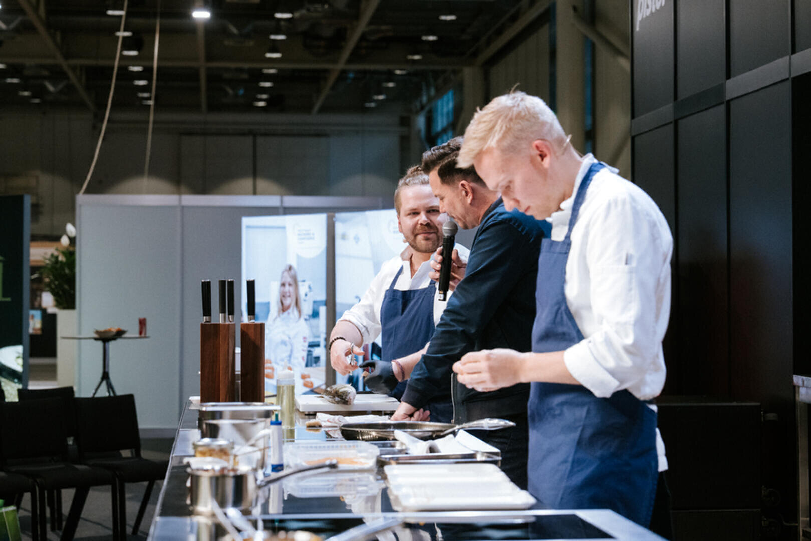 Drei Köche in blauen Schürzen stehen an einer Theke und bereiten bei einer Kochvorführung in einem modernen Innenraum Speisen zu. Ein Koch spricht, während die anderen sich auf das Anrichten der Gerichte konzentrieren.