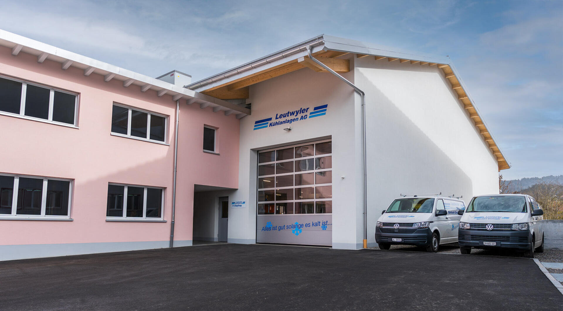 Ein modernes Industriegebäude mit rosa-weißem Äußeren, großen Fenstern und einem gläsernen Garagentor. Zwei weiße Lieferwagen sind auf einer gepflasterten Auffahrt geparkt. Der Himmel ist teilweise bewölkt.