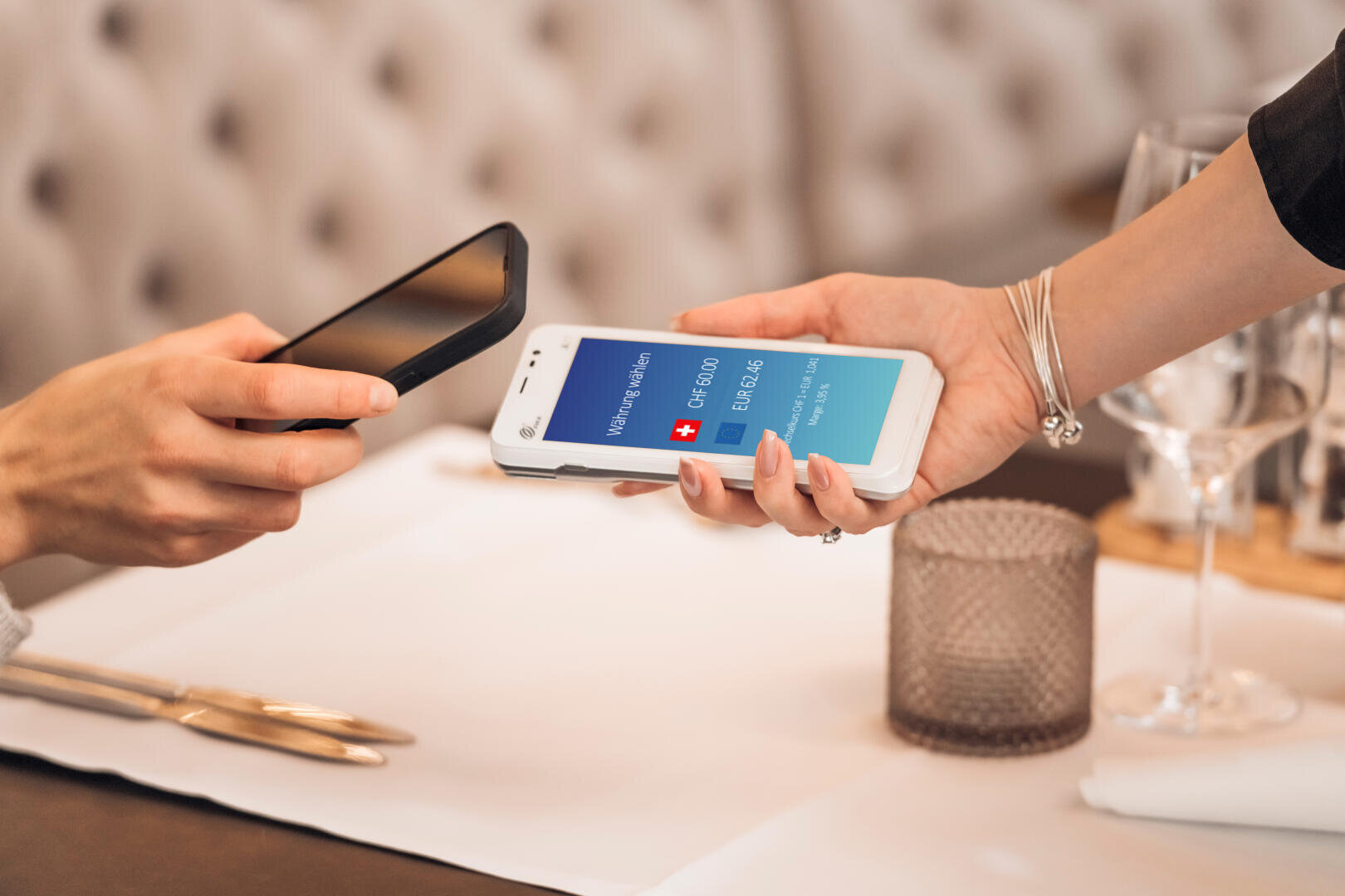 Deux personnes échangent un paiement numérique dans un restaurant, à l'aide de smartphones. L'une des personnes tient un téléphone affichant une application de paiement suisse, tandis que l'autre le scanne avec son appareil au-dessus d'une table.