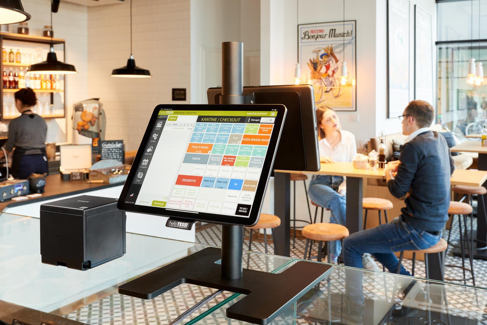 Ein modernes Kassensystem mit Touchscreen steht auf einem Tresen in einem hellen Café. Im Hintergrund sitzen Kunden an Tischen und hinter dem Tresen arbeitet eine Bedienung.