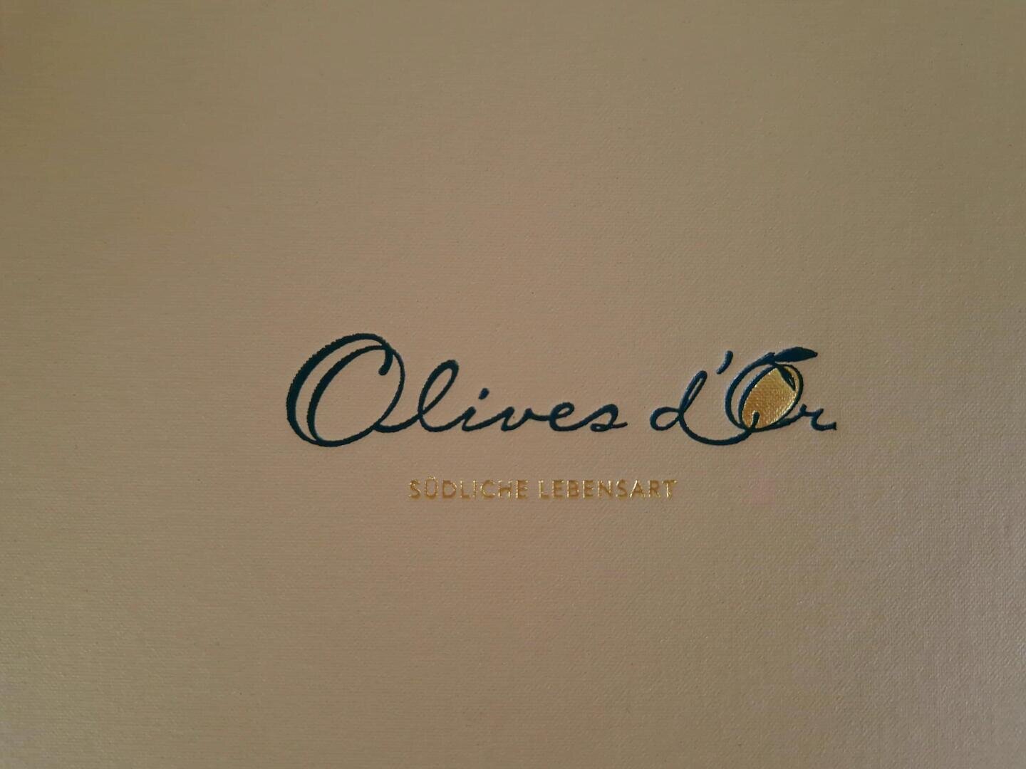 Beigefarbener Hintergrund mit eleganter grüner und goldener Schrift "Olives dOr" und kleinerer goldener Schrift darunter "Südliche Lebensart".