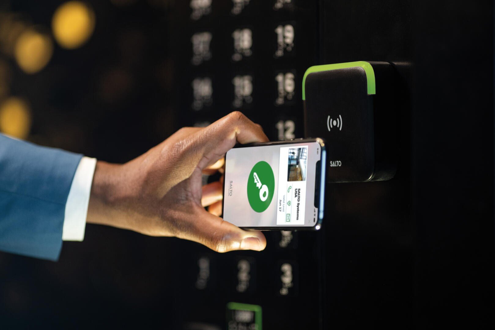 Eine Person hält ein Smartphone, das einen digitalen Schlüssel anzeigt, in die Nähe eines schwarzen elektronischen Zugangslesers und ist im Begriff, eine Tür mithilfe der mobilen Zugangstechnologie zu entsperren.