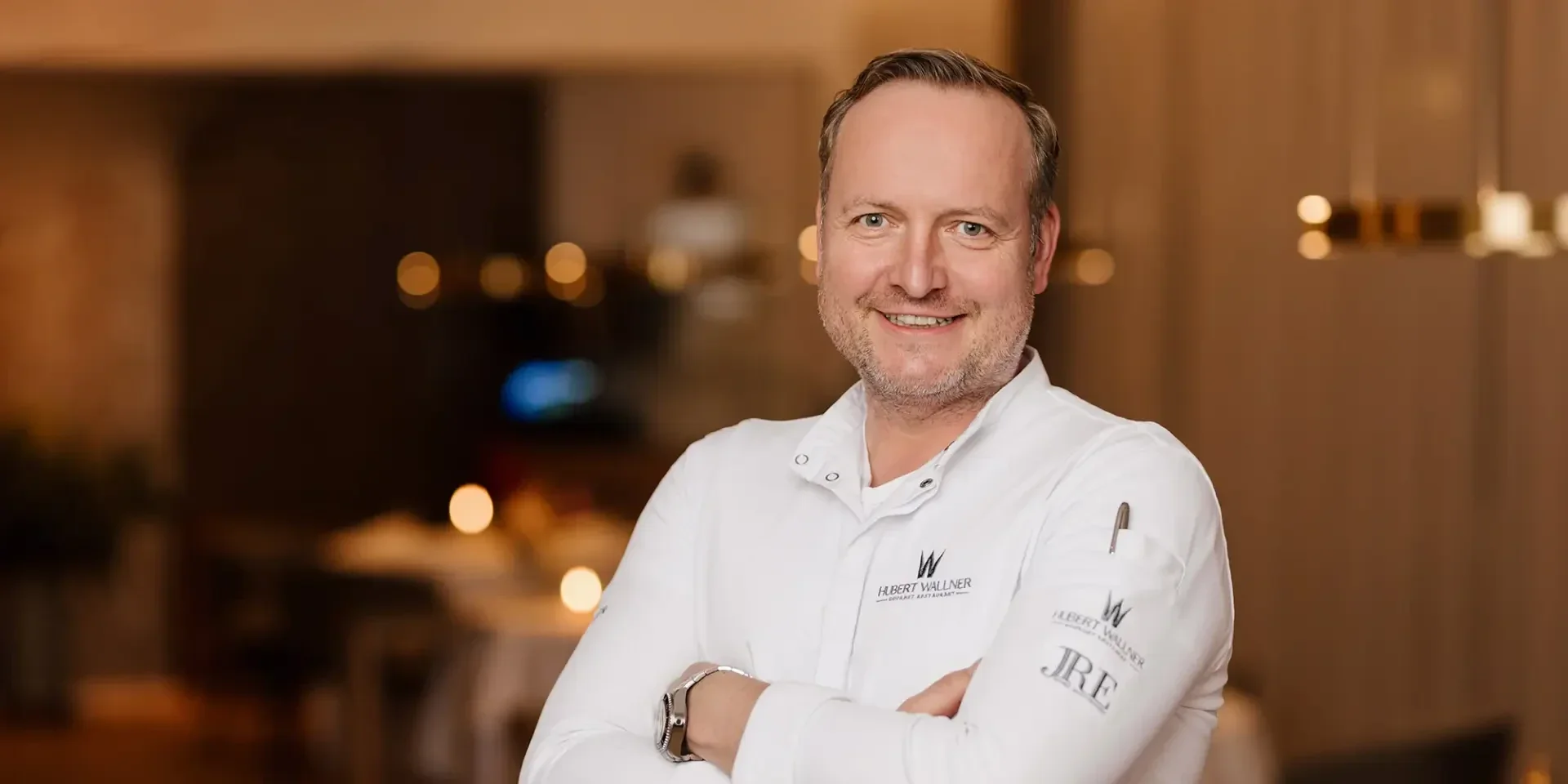 Un cuisinier souriant aux cheveux clairs, vêtu d'une veste de cuisine blanche, se tient les bras croisés dans un restaurant chaleureusement éclairé. L'arrière-plan est légèrement flou, ce qui souligne l'atmosphère accueillante.