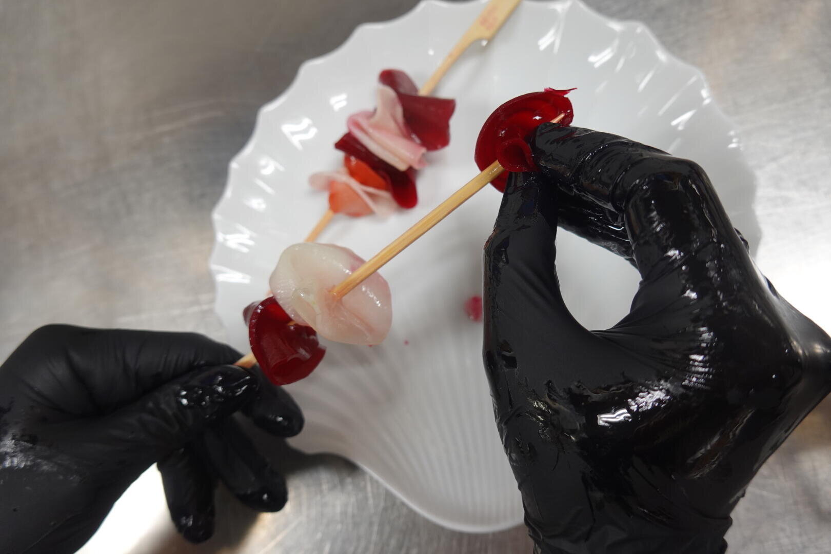Deux mains portant des gants noirs tiennent une brochette avec des morceaux rouges et translucides, peut-être des bonbons ou de la gelée, au-dessus d'une assiette blanche festonnée sur une surface métallique.