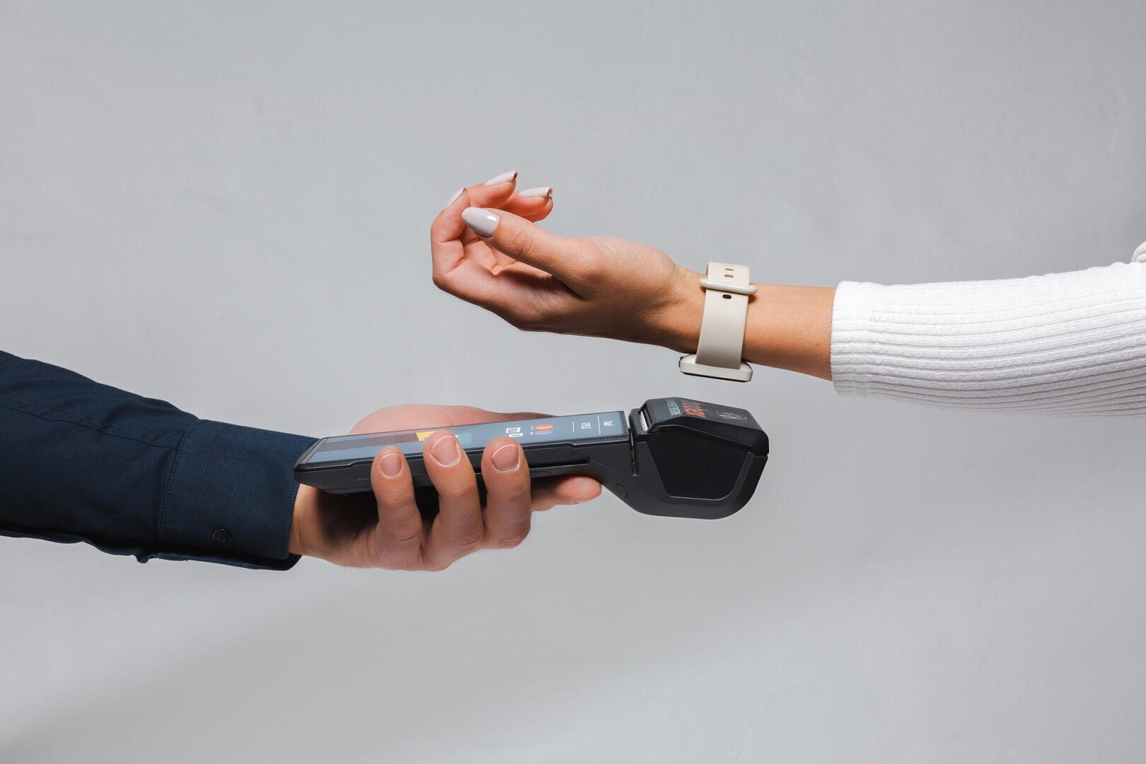 Eine Person, die eine intelligente Uhr trägt, hält ihr Handgelenk in die Nähe eines drahtlosen Zahlungsterminals, das von einer anderen Person gehalten wird, und demonstriert so das kontaktlose Bezahlen.