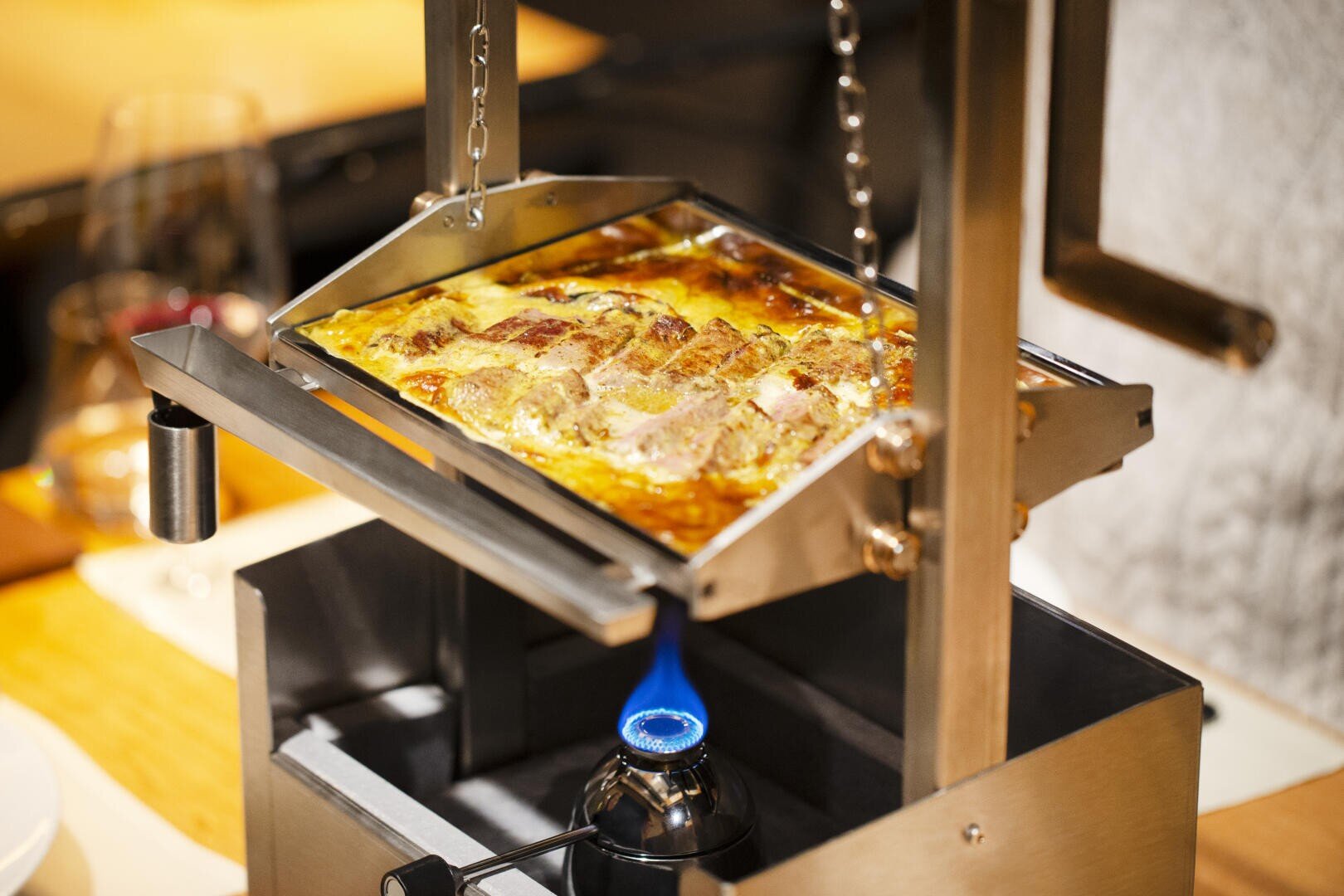 Ein Raclette-Grill aus Metall mit einer Flamme an der Unterseite schmilzt sanft ein Tablett mit einer Käseplatte, wodurch eine goldene, blubbernde Oberfläche entsteht. Die Einrichtung steht auf einem Holztisch, im Hintergrund sind unscharfe Gläser zu sehen.