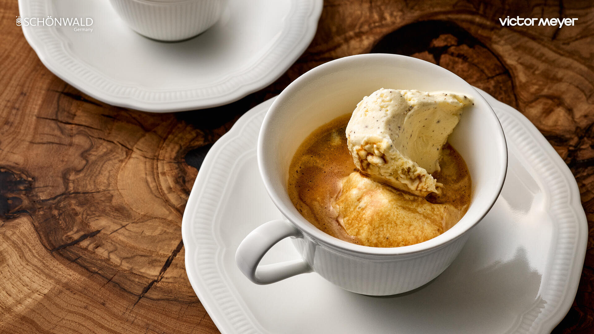Eine weiße Tasse, gefüllt mit Espresso und einer Kugel Vanilleeis, steht auf einer weißen Untertasse auf einem Holztisch. Eine weitere Tasse und Untertasse sind teilweise im Hintergrund zu sehen.
