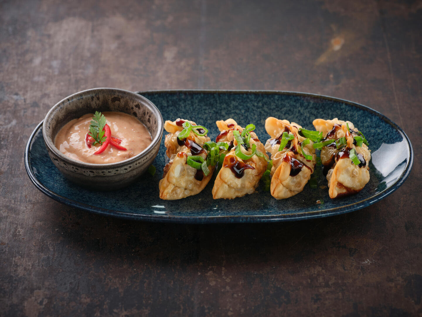 Vier gebratene Teigtaschen mit gehackten grünen Zwiebeln und dunkler Soße werden auf einem dunklen ovalen Teller angerichtet und mit einer Schale Dip-Soße, garniert mit Koriander und roten Chilischeiben, serviert.