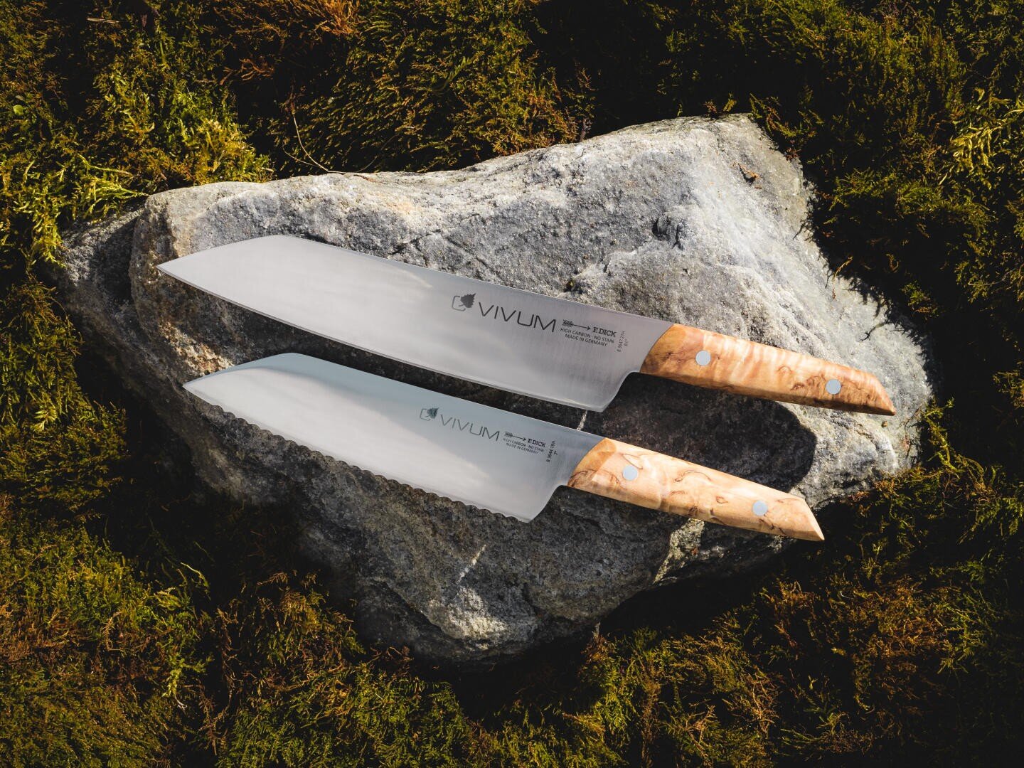 Deux couteaux de cuisine aux manches en bois clair et aux lames brillantes reposent sur un gros rocher gris, entouré de mousse verte. Les couteaux sont positionnés parallèlement l'un à l'autre, le nom de la marque Vivum étant visible sur les deux lames.