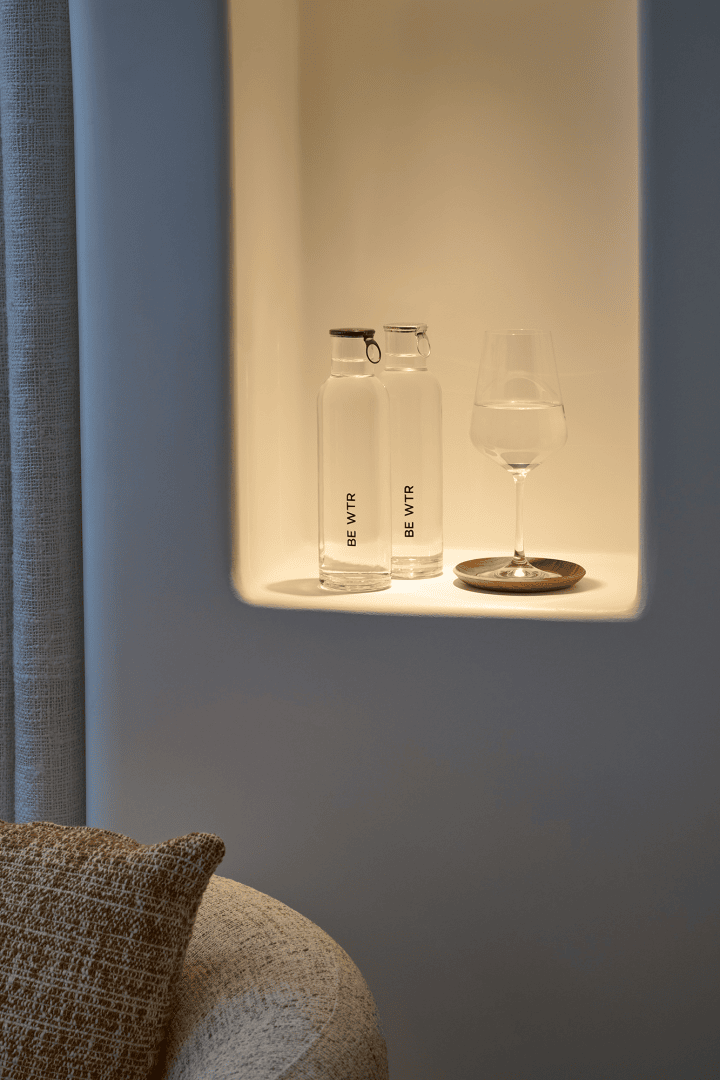 Deux bouteilles en verre portant l'inscription BEM VYR et un verre à vin sur un dessous de plat en bois sont placés dans une niche doucement éclairée, à côté d'un coussin beige structuré et d'un canapé clair incurvé.