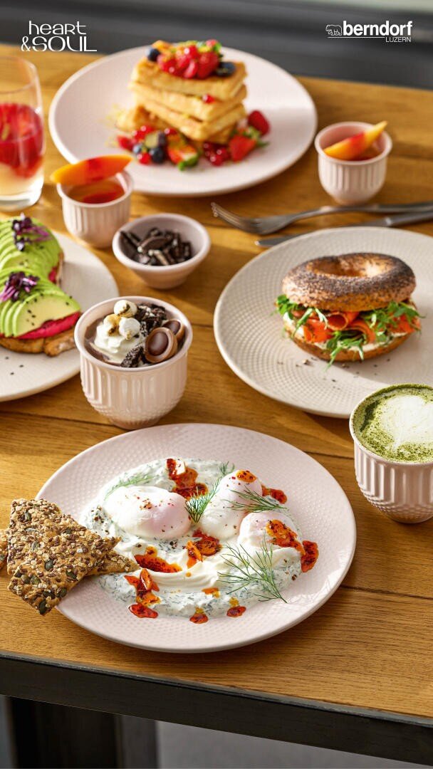 Ein Holztisch, auf dem pochierte Eier auf Joghurt mit Crackern, ein Bagel mit Gemüse, Waffeln mit Beeren, Avocado-Toast, kleine Schälchen mit Snacks, Matcha Latte und Getränke angerichtet sind. Oben erscheinen die Logos heart & SOUL und berndorf.