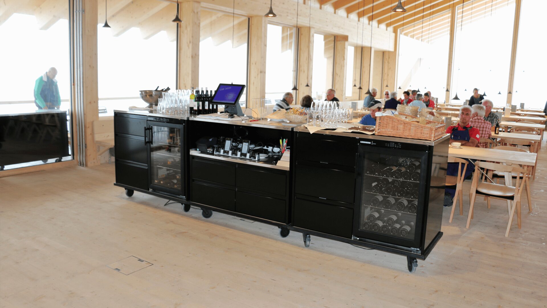 Une station de self-service avec caisse enregistreuse, verres, couverts et bouteilles de vin se trouve dans une salle à manger en bois, lumineuse et moderne, où les clients sont assis à des tables.
