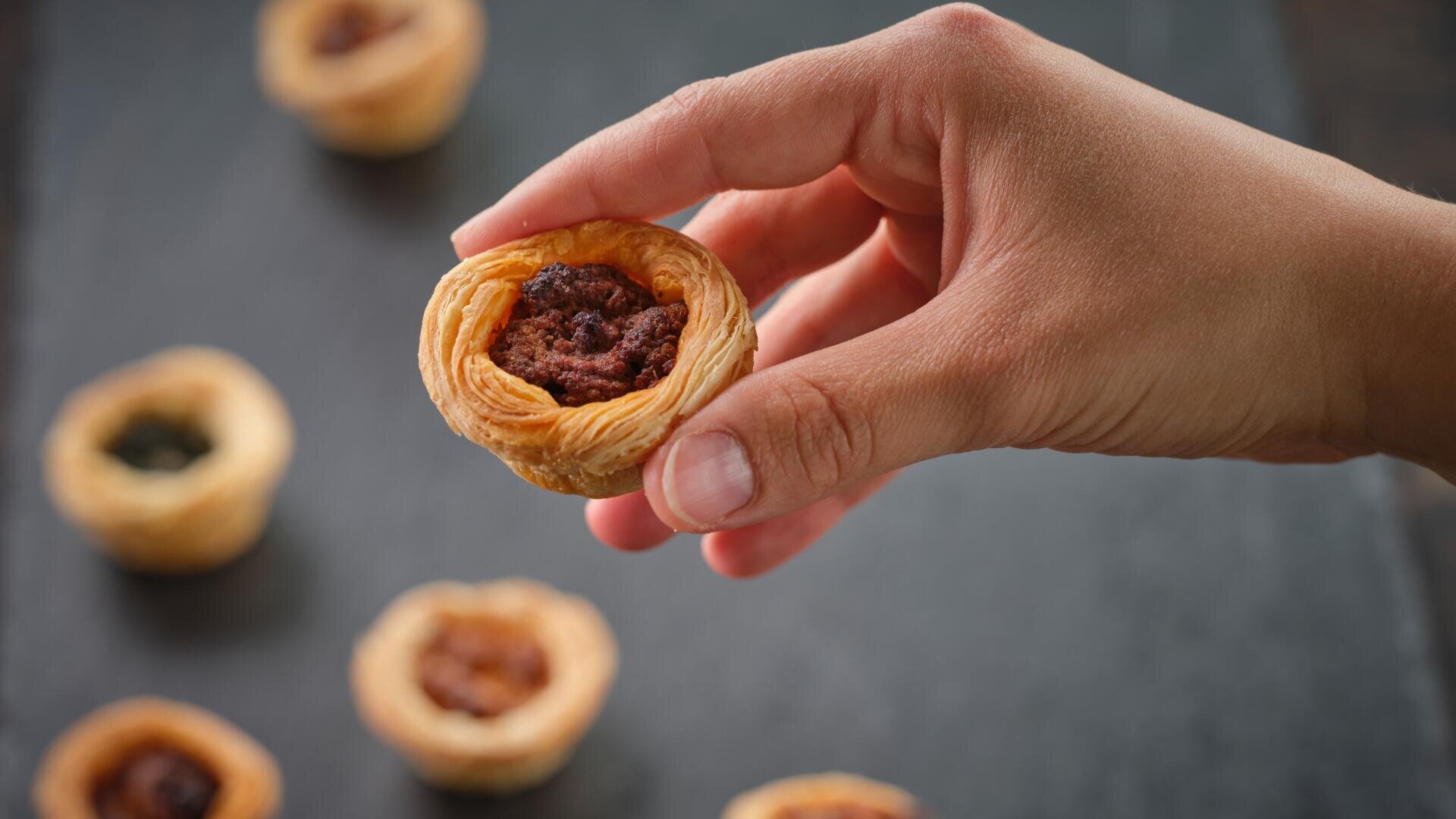 Eine Hand hält ein kleines, rundes Gebäck mit einer flockigen Kruste und einer dunklen Füllung; mehrere ähnliche Gebäcke sind im Hintergrund auf einer dunklen Fläche verschwommen zu erkennen.