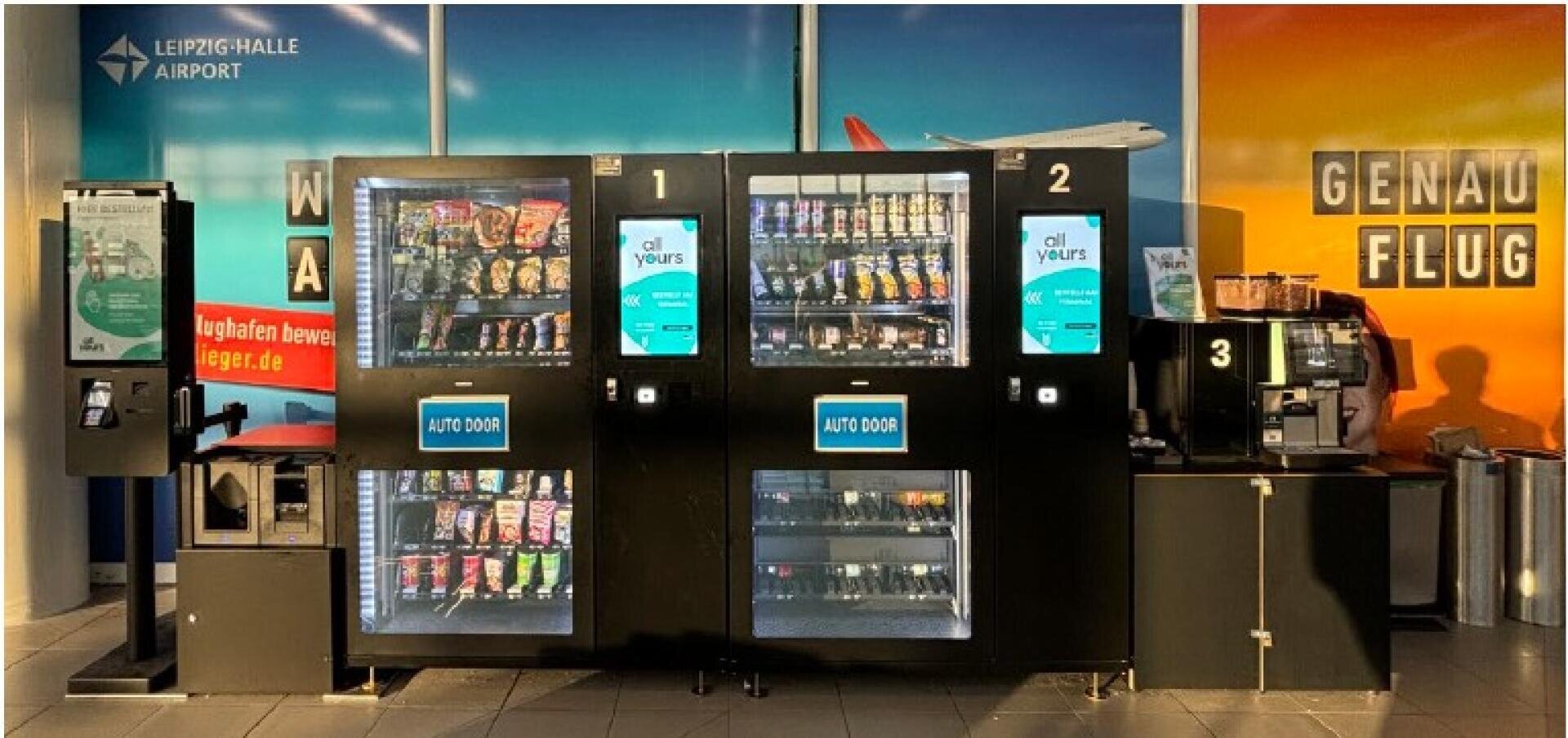 Am Flughafen Leipzig-Halle sind drei Automaten aufgereiht, die Snacks, Getränke und Kaffee anbieten. Die Automaten sind nummeriert und stehen an einer bunten Wand mit Schildern in Deutsch und Englisch.