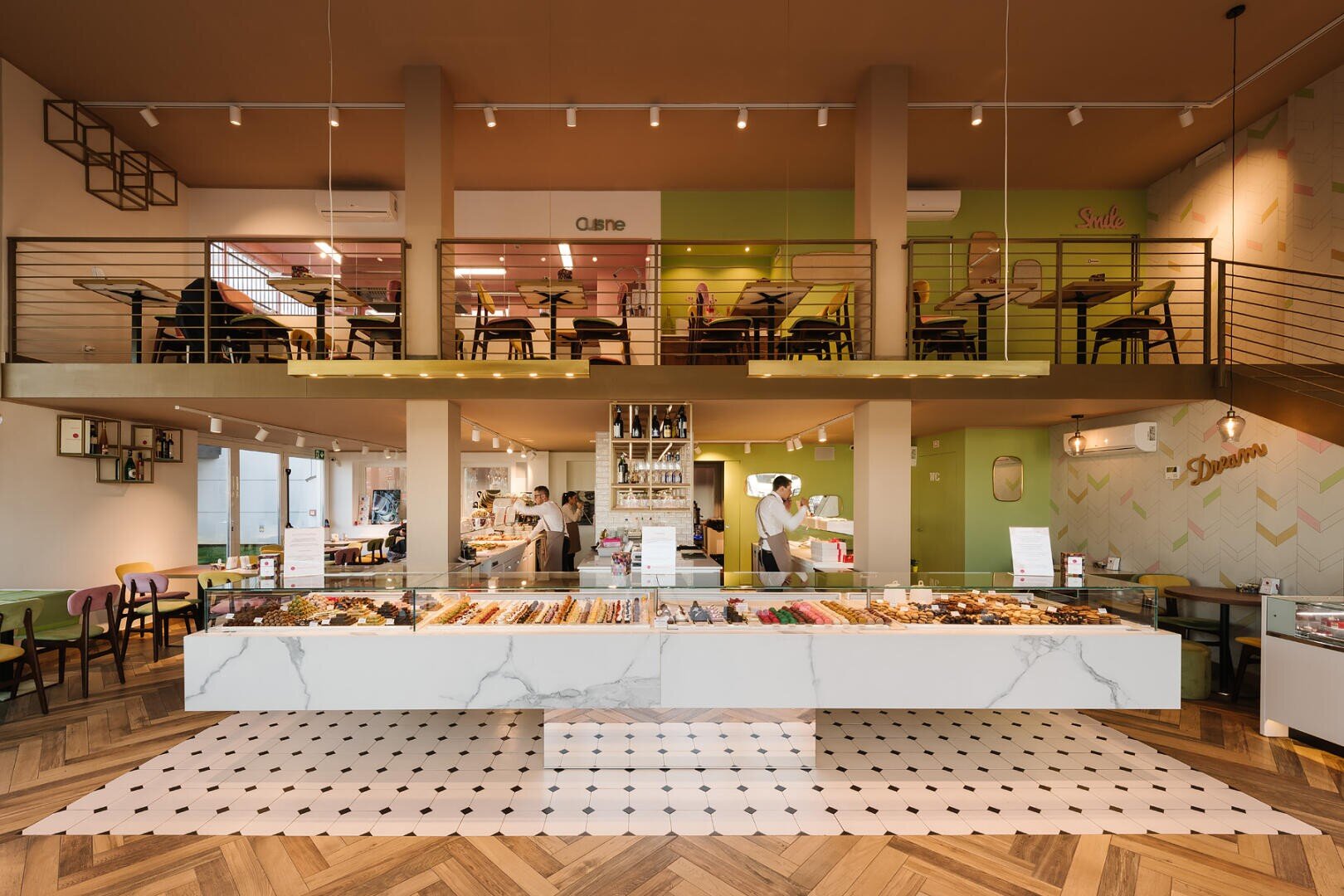Eine moderne, geräumige Bäckerei mit einer großen Vitrine in der Mitte, die mit Backwaren gefüllt ist, luftigen Sitzgelegenheiten im Erdgeschoss und einem Zwischengeschoss im Obergeschoss, warmer Beleuchtung und einer stilvollen Einrichtung in Grün- und Beigetönen.