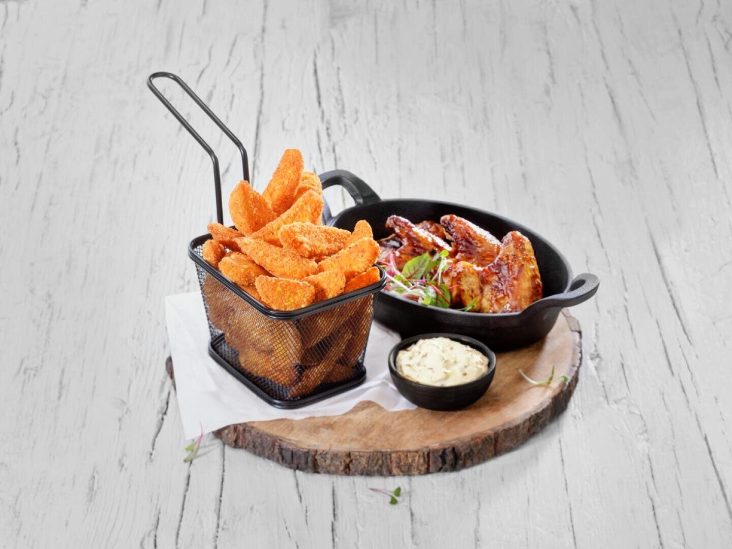 Une portion de pommes de terre assaisonnées dans un panier métallique, des ailes de poulet grillées dans un petit plat en fonte et un bol de sauce, le tout disposé sur une planche en bois rustique.