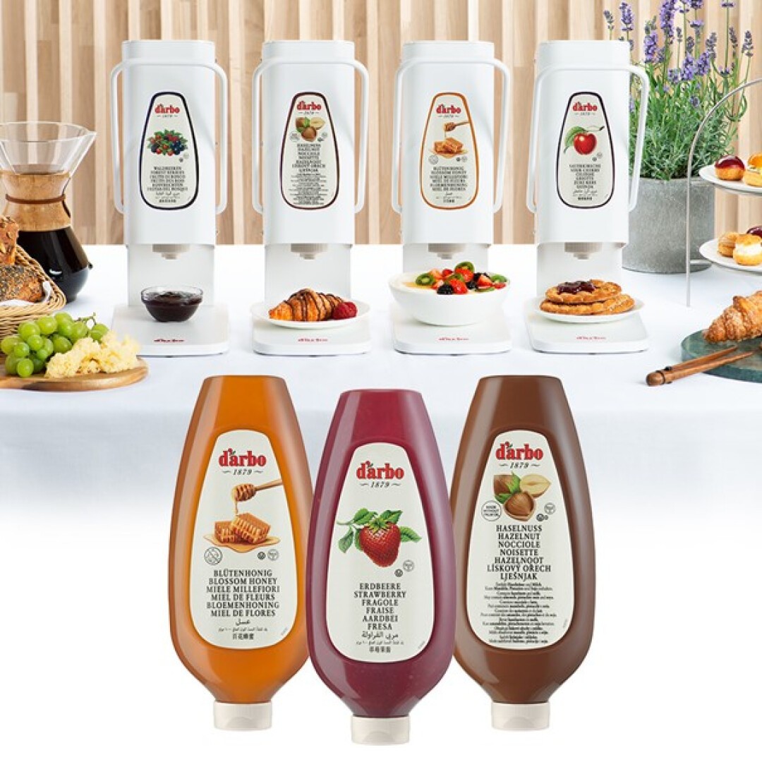 Ein Frühstücksbuffet mit Gebäck, Obst und Brot vor drei Spendern für weißen Sirup und drei Flaschen D’Arbo-Saucen: Honig, Erdbeere und Schokoladen-Haselnuss.