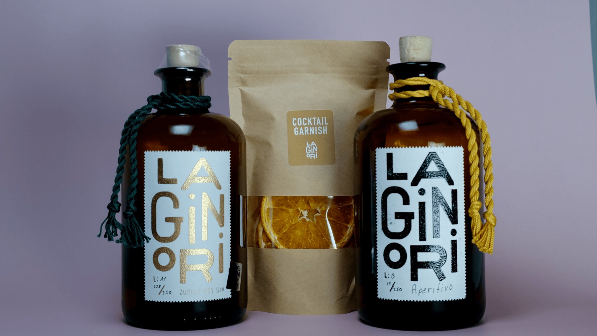 Deux bouteilles en verre marron étiquetées LA GINORI avec des cordons décoratifs, l'un vert et l'autre jaune, se trouvent à côté d'un sachet marron étiqueté Cocktail Garnish contenant des tranches d'orange séchées, le tout sur un fond violet clair.