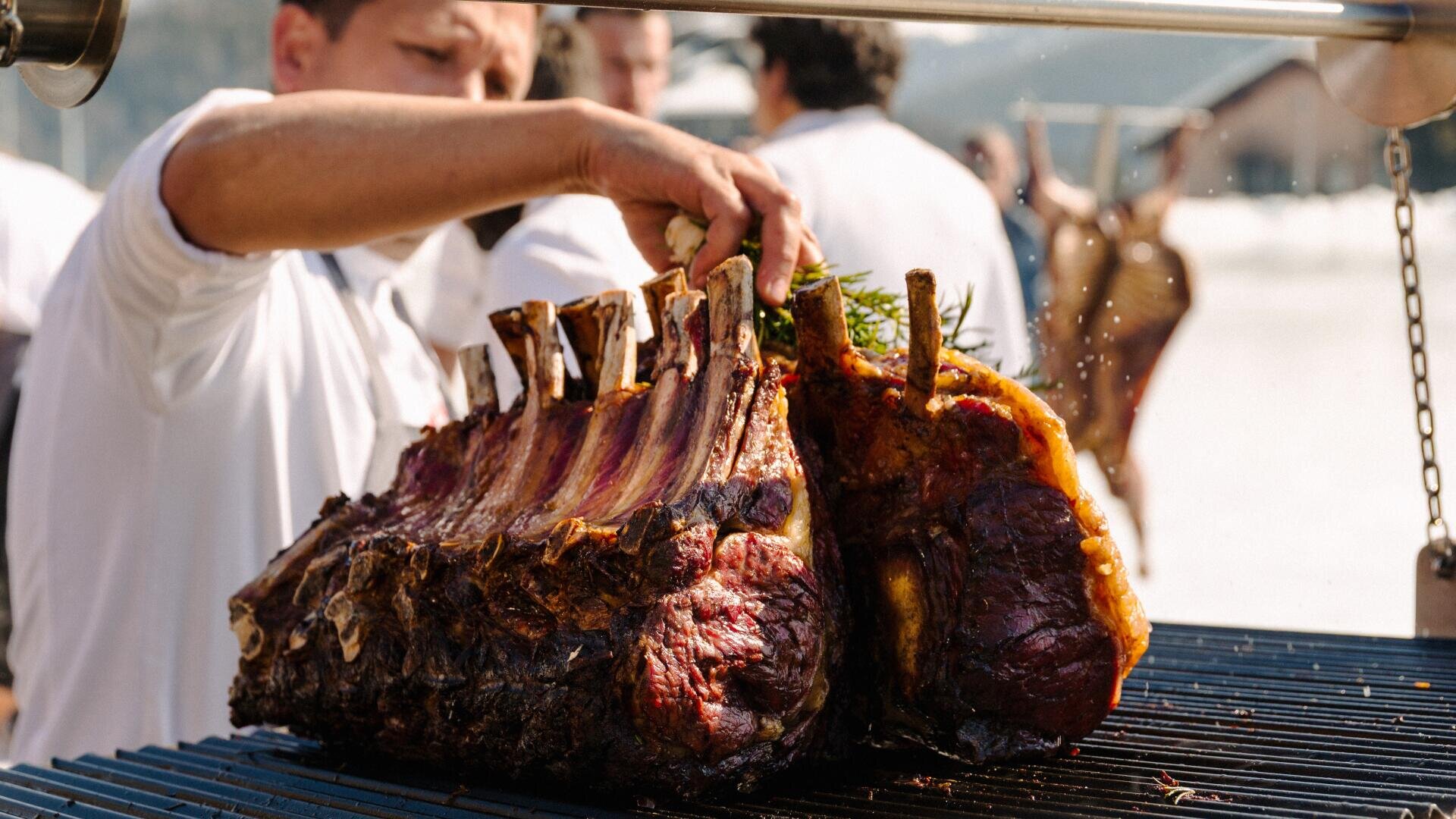 Ein Koch im weißen Kittel grillt im Freien große Rinderrippchen und kümmert sich sorgfältig um das Fleisch, während es über einer offenen Flamme gart, während im Hintergrund andere Menschen und eine verschwommene Landschaft zu sehen sind.