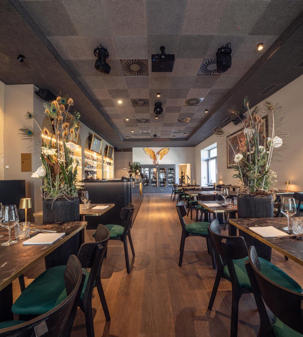 Modernes Restaurant mit Holzböden, Tischen mit Gläsern, grünen und schwarzen Stühlen, großen Blumen, einer stilvollen Bar und einer goldenen Schmetterlingswand im hinteren Bereich. Die warme Beleuchtung schafft eine gemütliche Atmosphäre.