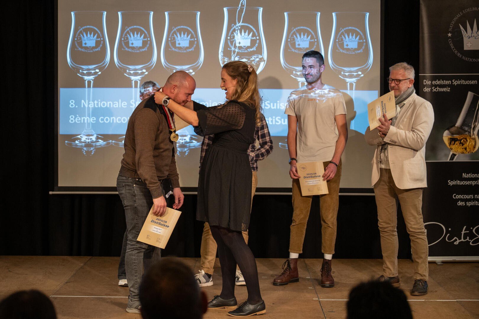Eine Frau legt einem lächelnden Mann auf der Bühne eine Medaille um den Hals. Zwei Männer stehen in der Nähe und halten Urkunden in der Hand, während ein weiterer Mann zusieht. Auf der Leinwand hinter ihnen werden Grafiken mit Auszeichnungen projiziert.