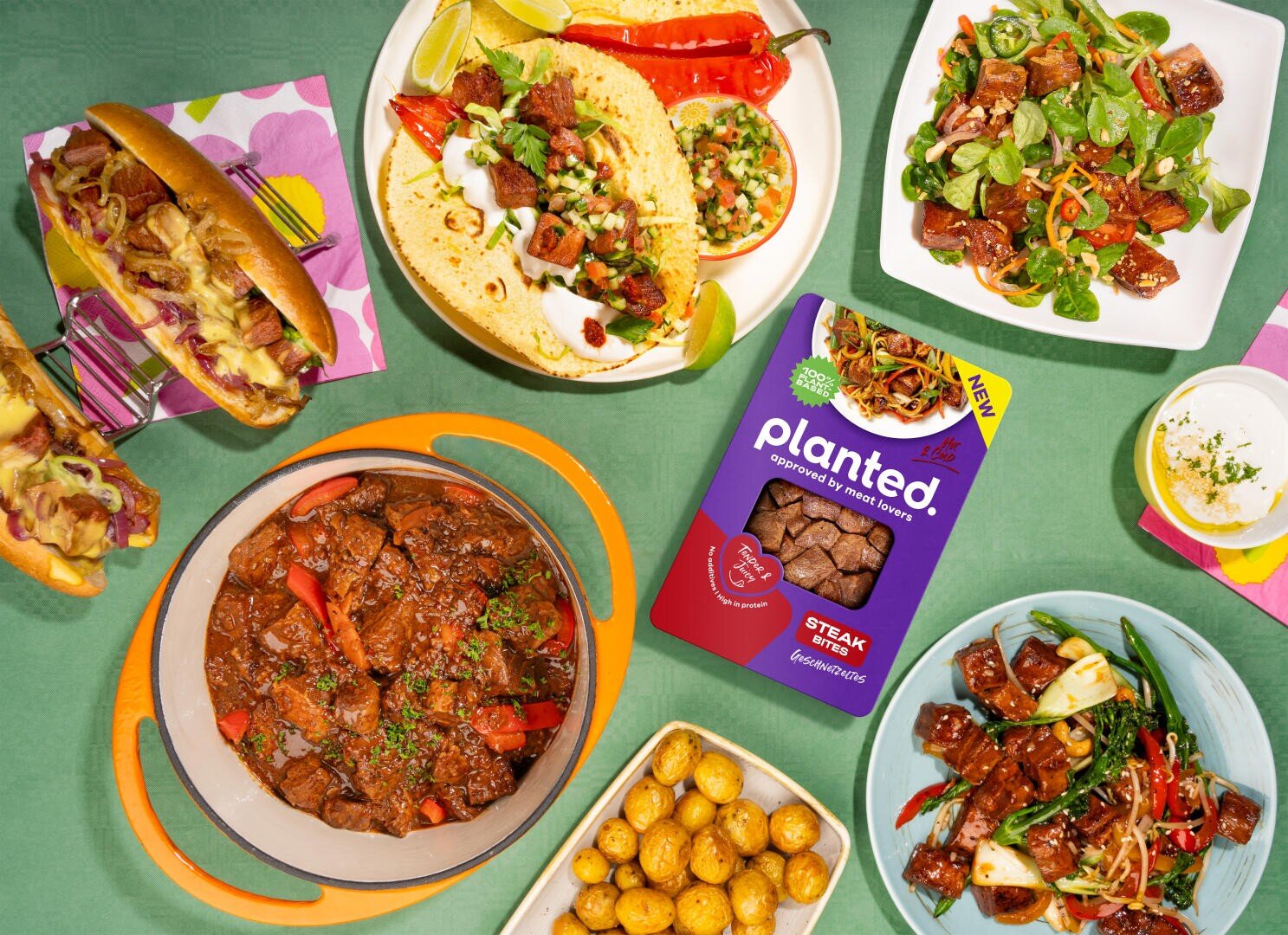 Une table colorée avec différents plats préparés avec du steak végétal, dont des tacos, un ragoût, une salade, un hot dog, des brochettes, des pommes de terre sautées et une boîte contenant le produit végétal.