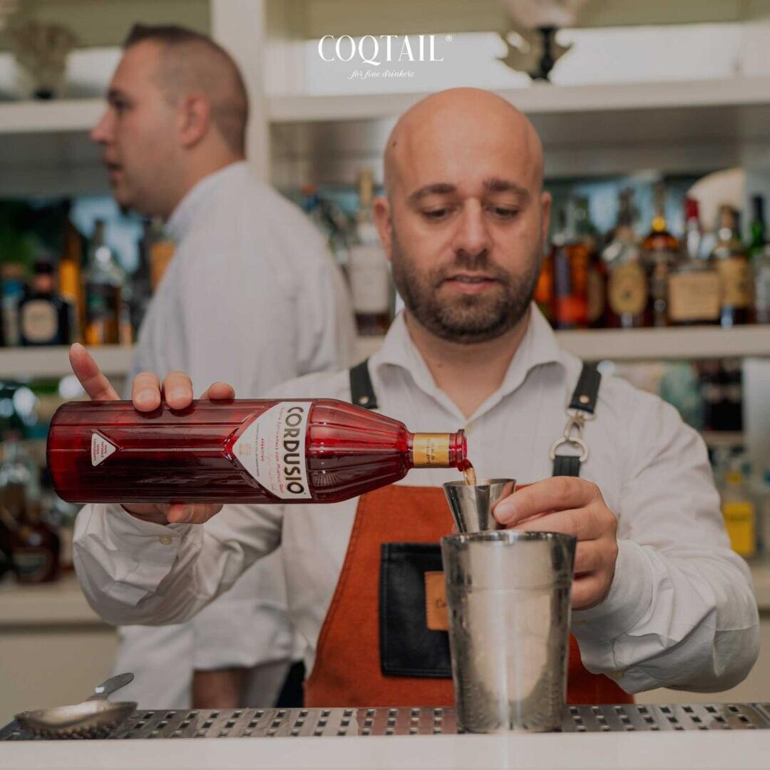 Ein Barkeeper in weißem Hemd und brauner Schürze gießt roten Likör in einen Metallstreuer, hinter ihm stehen Alkoholflaschen in Regalen. Eine weitere Person steht im Hintergrund. Oben ist das Wort COQTAIL zu sehen.