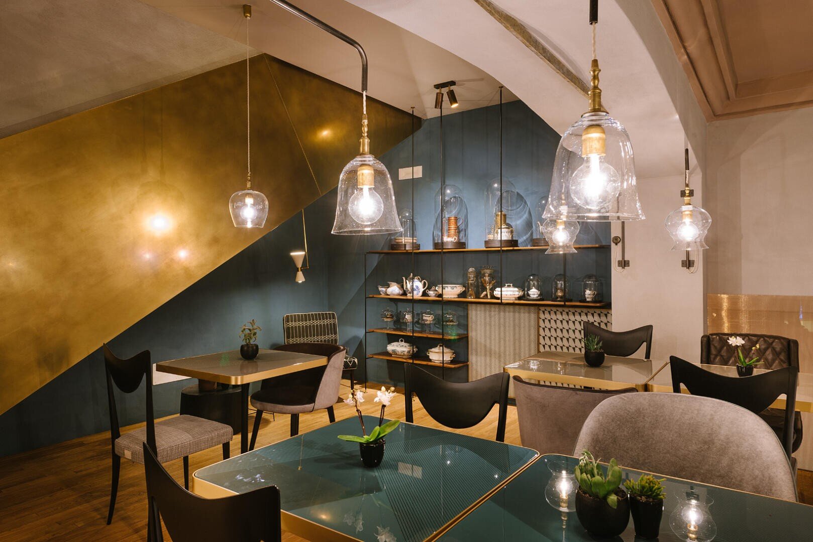 Un intérieur de café moderne avec des lampes suspendues en verre, des chaises élégantes, de petites plantes en pot sur des tables en verre, et un mur décoratif avec des étagères contenant des théières, des tasses et des ornements. L'éclairage est chaleureux et accueillant.