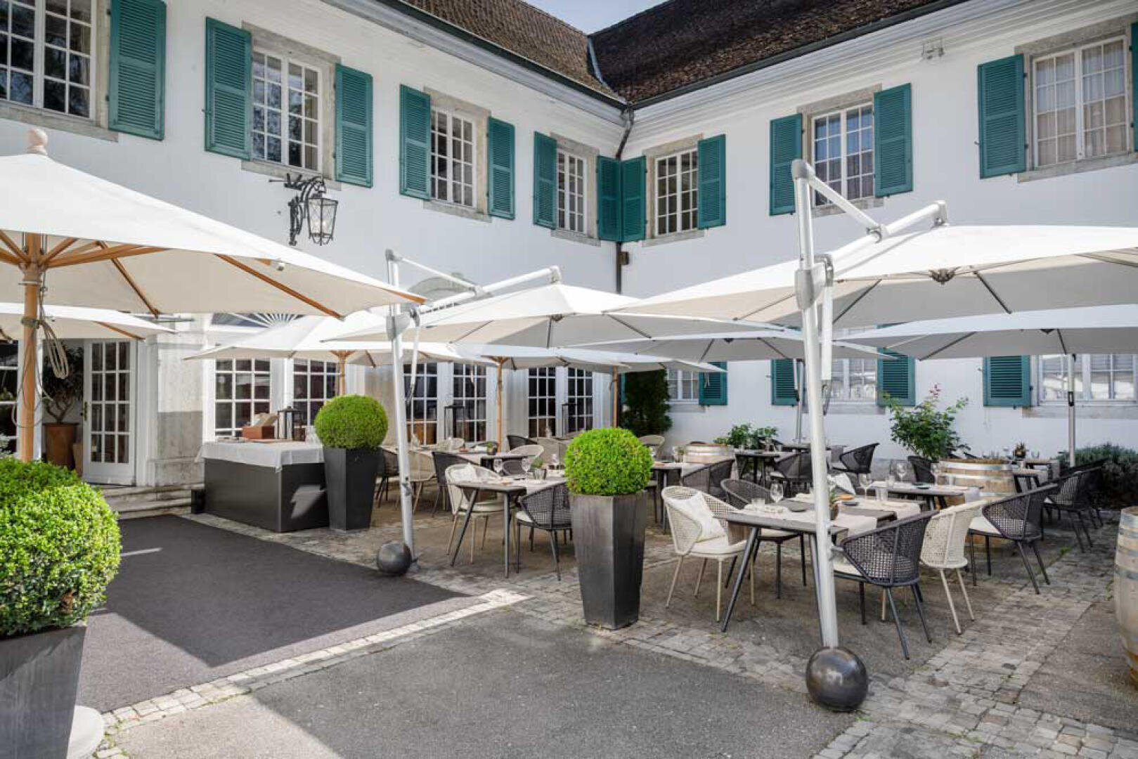 Restaurantterrasse mit Tischen und Stühlen unter großen weißen Sonnenschirmen, umgeben von Topfpflanzen, vor einem weißen Gebäude mit türkisfarbenen Fensterläden und vielen Fenstern.