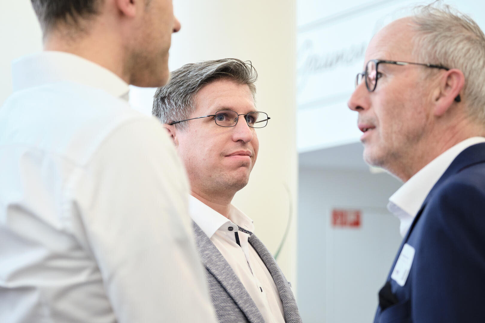 Trois hommes en tenue d'affaires discutent à l'intérieur. L'un d'entre eux porte des lunettes et un blazer gris clair, tandis que les deux autres sont partiellement visibles.