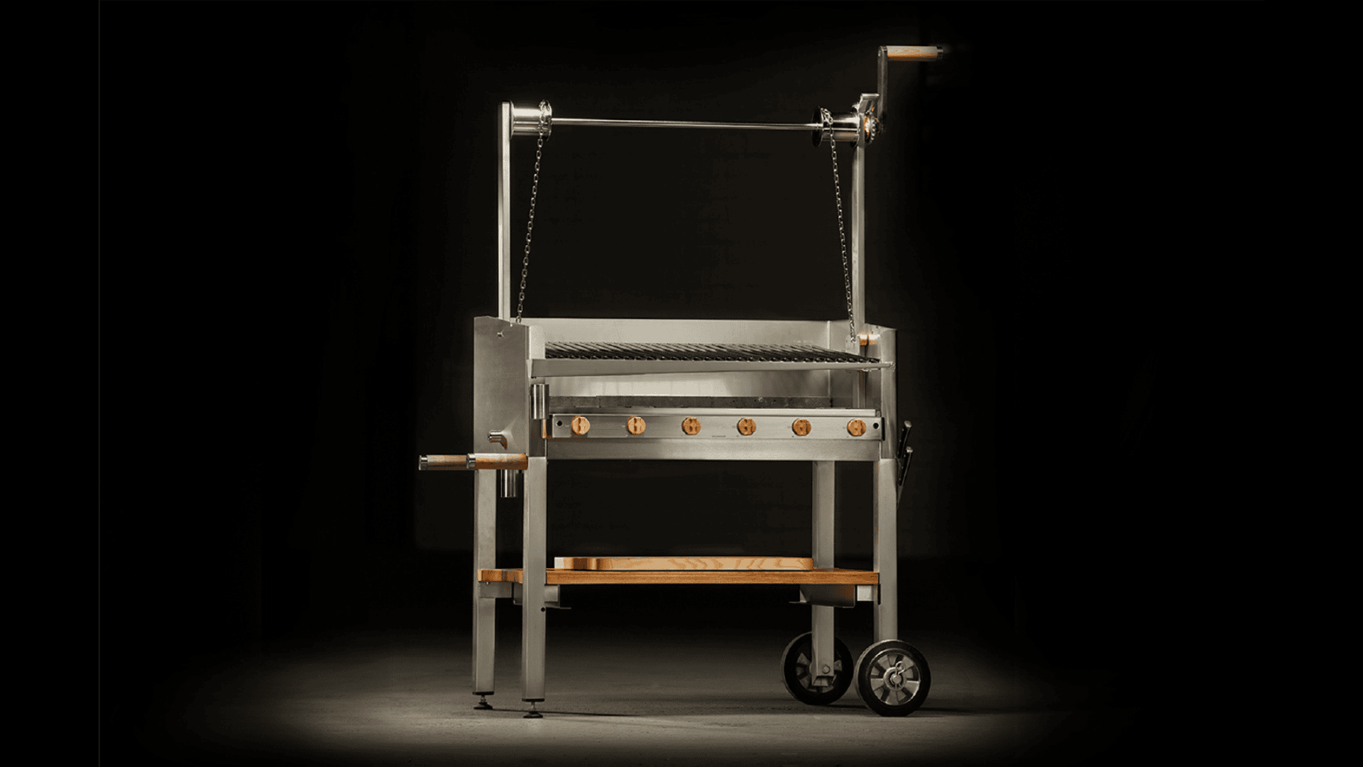 Ein moderner Edelstahlgrill für den Außenbereich mit Holzakzenten, einer Kurbel zum Einstellen der Grillhöhe und zwei Rädern für die Mobilität, dargestellt vor einem dunklen Hintergrund.