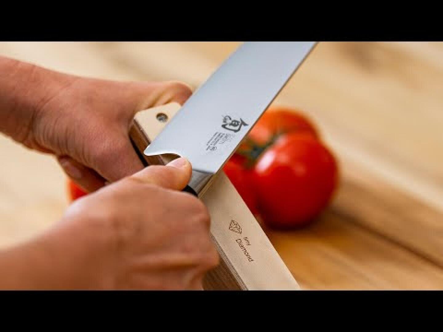 Une personne aiguise un couteau de cuisine à l'aide d'une raclette sur une surface en bois, deux tomates mûres se confondant avec l'arrière-plan.