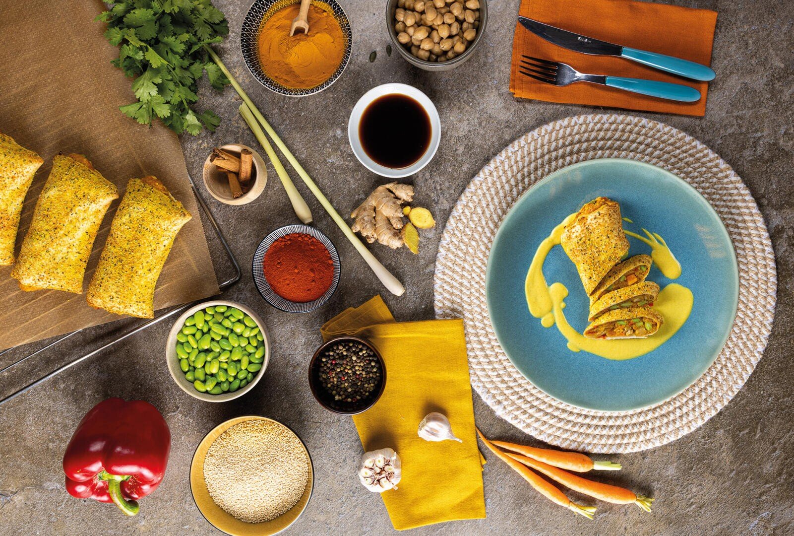 Eine farbenfrohe Draufsicht auf einen Tisch mit einem gefüllten Wrap auf gelber Soße, ungebackenen Wraps auf einem Tablett, Gewürzen, frischem Gemüse, Hülsenfrüchten, einer Paprika und Utensilien, die ordentlich auf einer strukturierten grauen Fläche angeordnet sind.