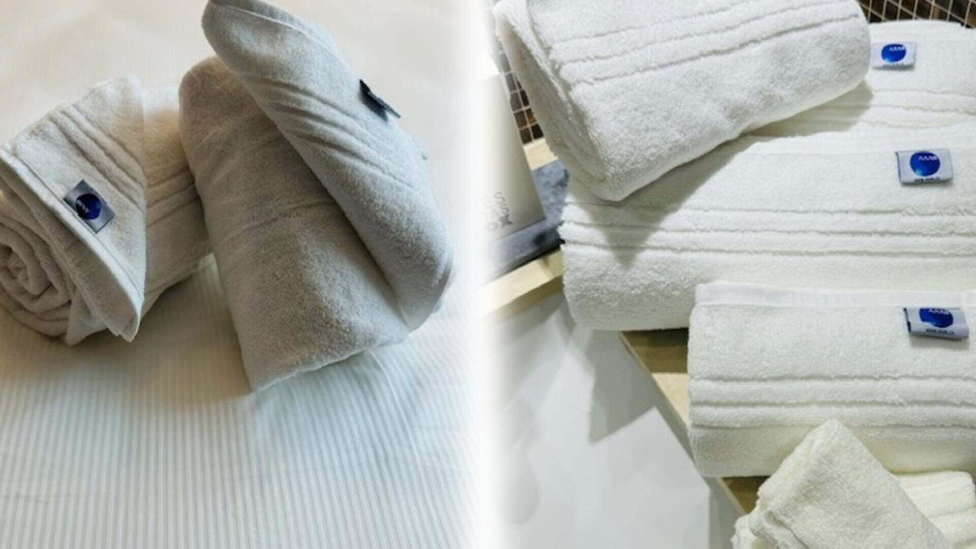 Deux ensembles de serviettes blanches bien pliées et enroulées sont posés sur des surfaces, chaque serviette est munie d'une petite étiquette bleue. L'ensemble de gauche est posé sur un lit blanc, celui de droite sur une étagère en bois.