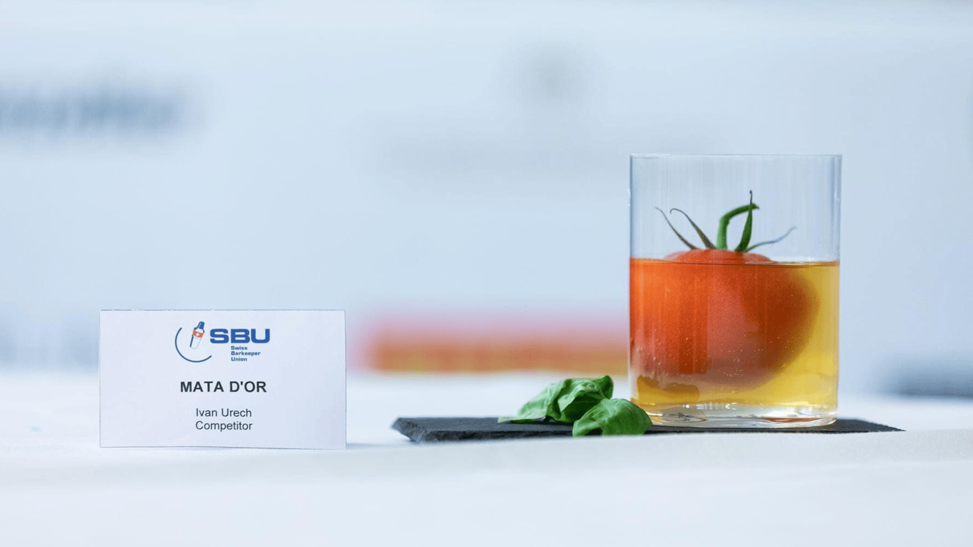 Un verre rempli d'un liquide clair et doré, dans lequel se trouve une tomate entière, est posé sur une ardoise à côté de feuilles de basilic. À côté du verre, on peut voir un badge avec l'inscription Mata Dor, Ivan Urech, Competitor.