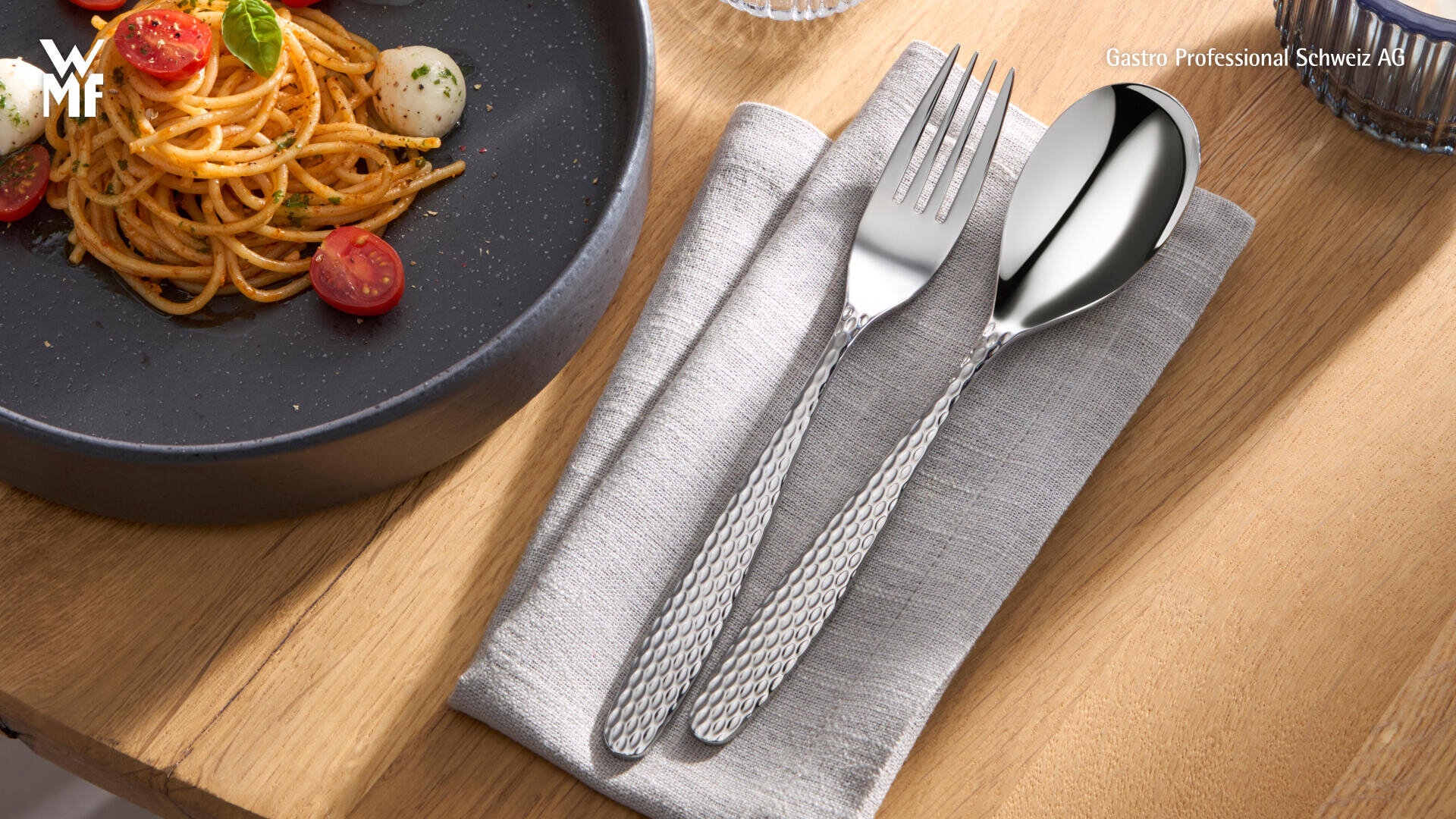 Un bol de spaghettis à la tomate est posé sur une table en bois à côté d'une serviette grise pliée sur laquelle sont posées une fourchette et une cuillère aux manches structurés.