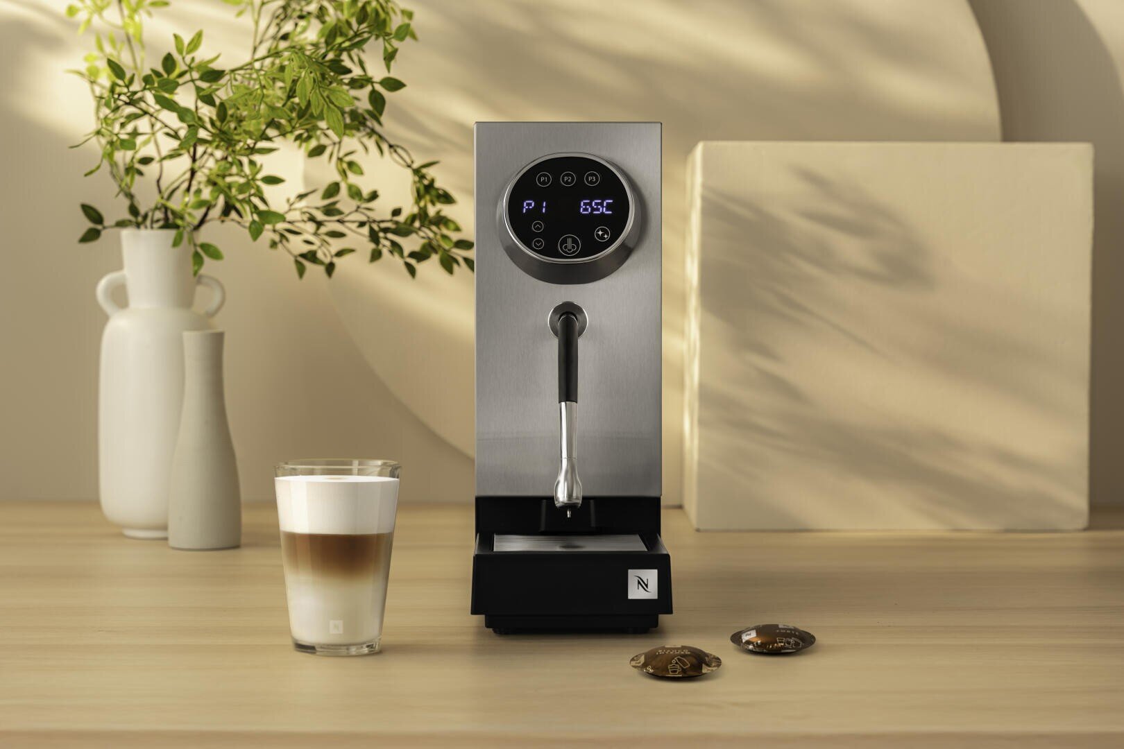 Eine moderne Espressomaschine mit Digitalanzeige steht auf einer hellen Holztischplatte, daneben ein Glas Milchkaffee und zwei Kaffeekapseln. Im Hintergrund ist eine weiße Vase mit grünen Zweigen zu sehen.