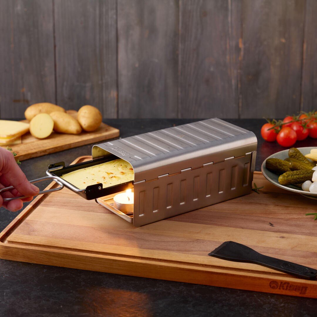 Ein Raclette-Grill aus Edelstahl mit einem Tablett mit schmelzendem Käse, der von einem Teelicht erhitzt wird, auf einem Holzbrett mit Kartoffeln, Tomaten und Gurken im Hintergrund.