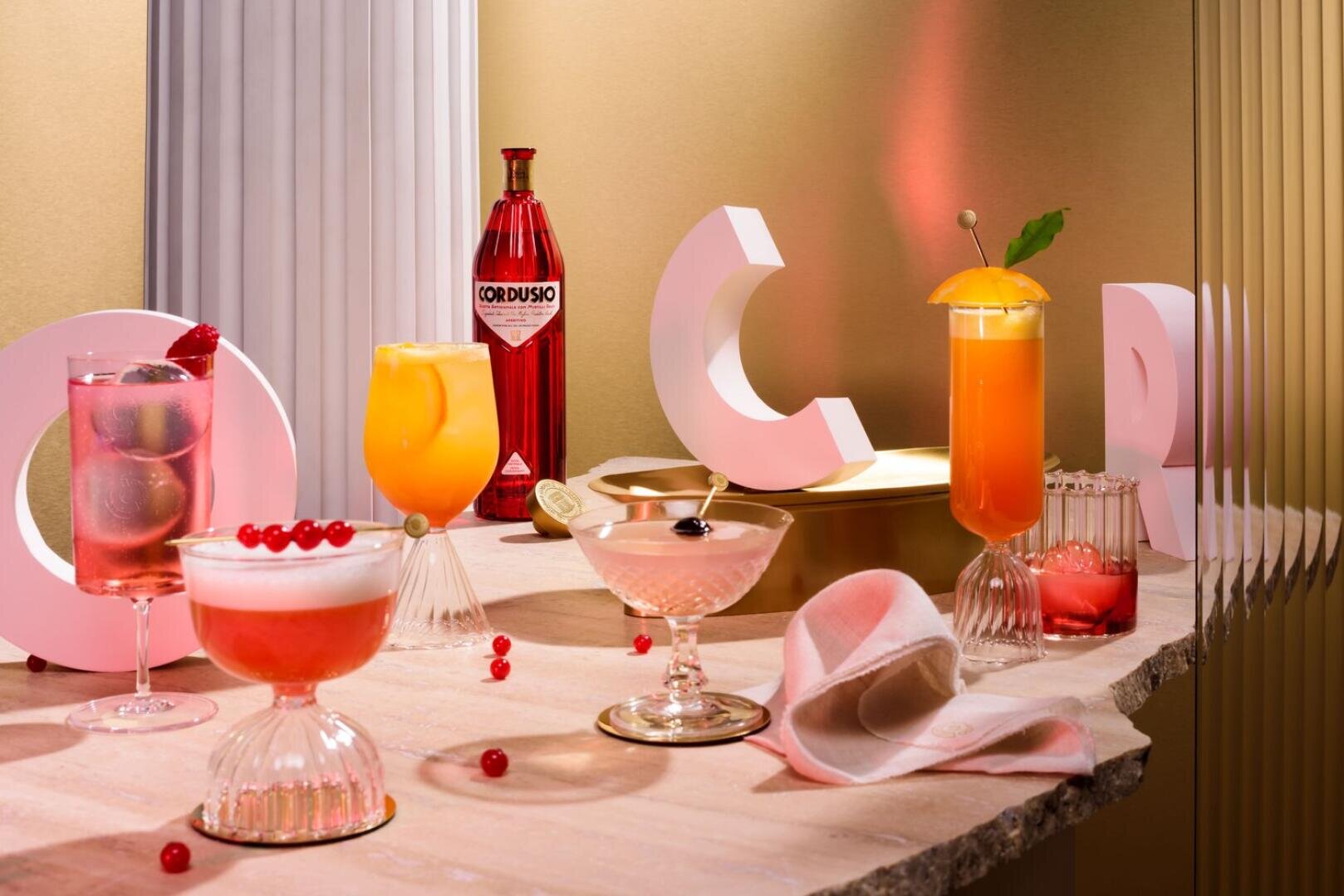 Verschiedene bunte Cocktails in eleganten Gläsern werden auf einer Steinfläche mit dekorativen geometrischen Stützen, einer roten Cordusio-Flasche, roten Johannisbeeren und einer gefalteten Serviette vor einem warmen, goldenen Hintergrund arrangiert.