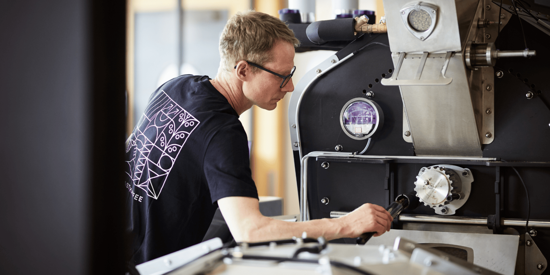 Dans une zone de travail bien éclairée, une personne portant des lunettes et un T-shirt noir règle un composant sur une grande machine industrielle, peut-être un torréfacteur de café.