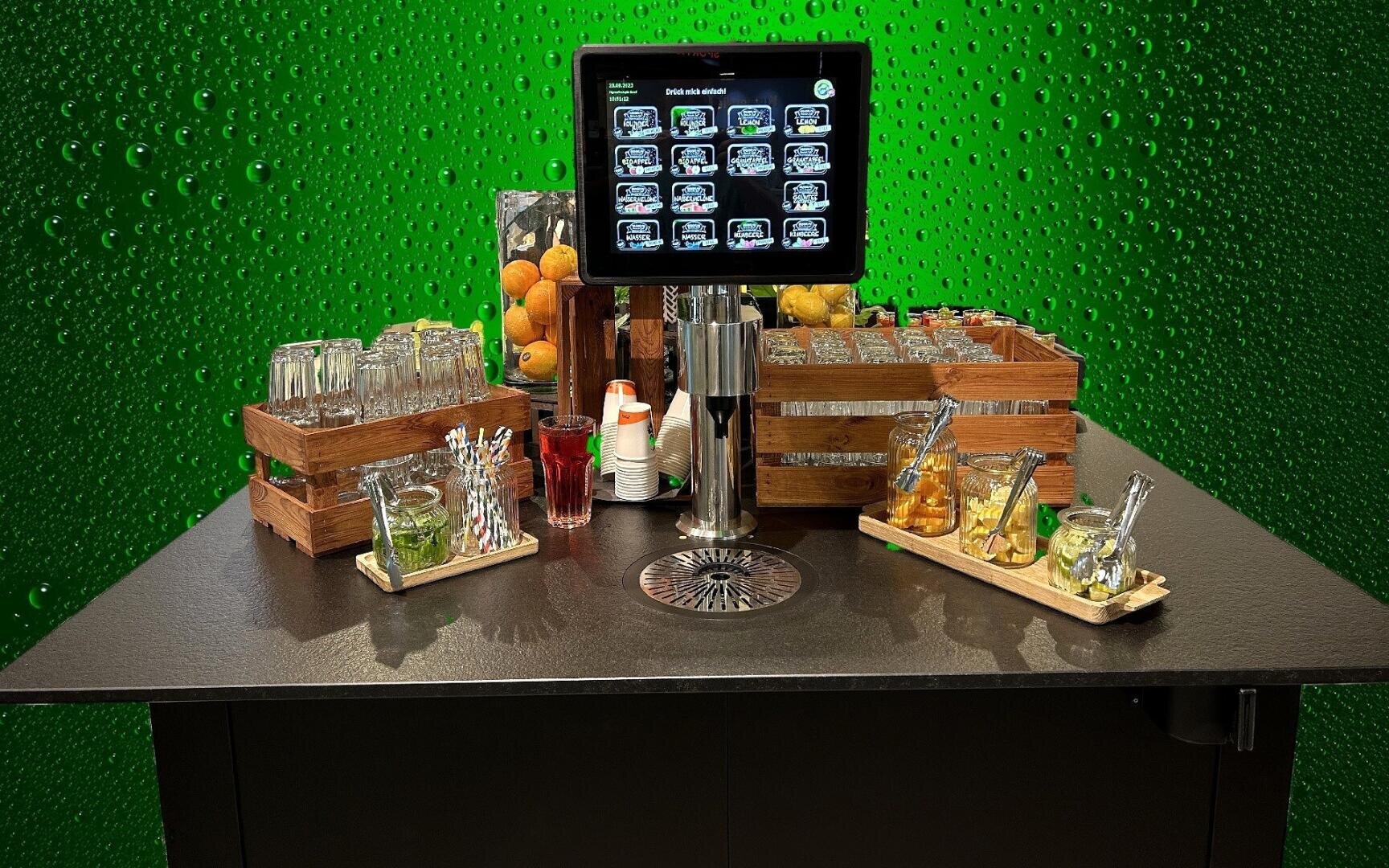 Eine Selbstbedienungs-Getränkestation mit digitalem Touchscreen, Gläsern, Fruchtgläsern, Zitronen, Limetten, Rührstäbchen und Utensilien auf einem schwarzen Tresen vor einem grünen, mit Wassertropfen gemusterten Hintergrund.